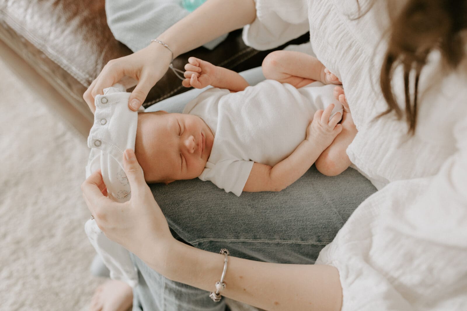 A mother sits at the edge of her couch and is about to stretch a white onesie over the head of her newborn. The baby is sleeping. The camera angle is from above looking down onto the mother's lap.