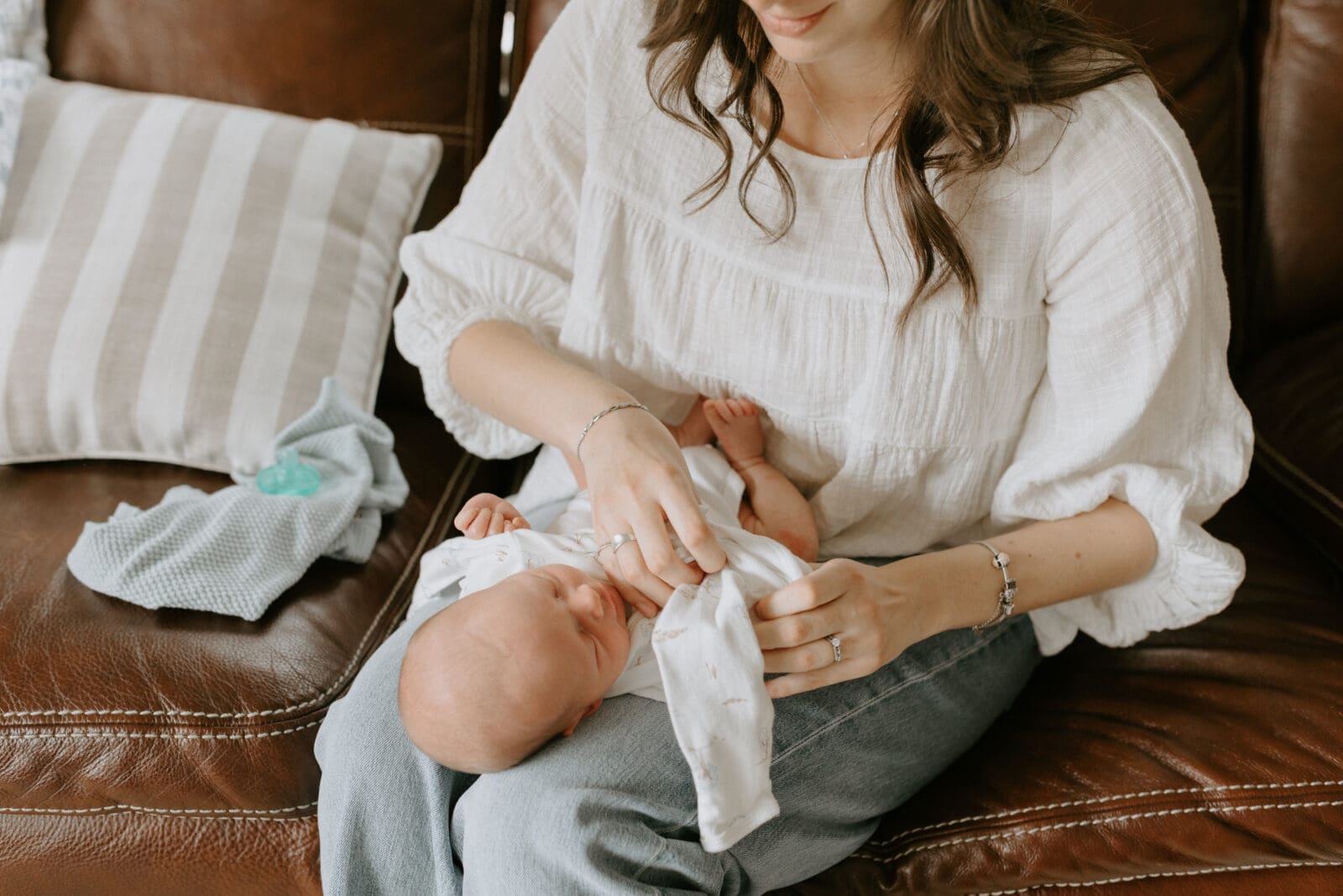 A mother sits on her brown couch with her baby on her lap. She is in the process of changing her baby's outfit.