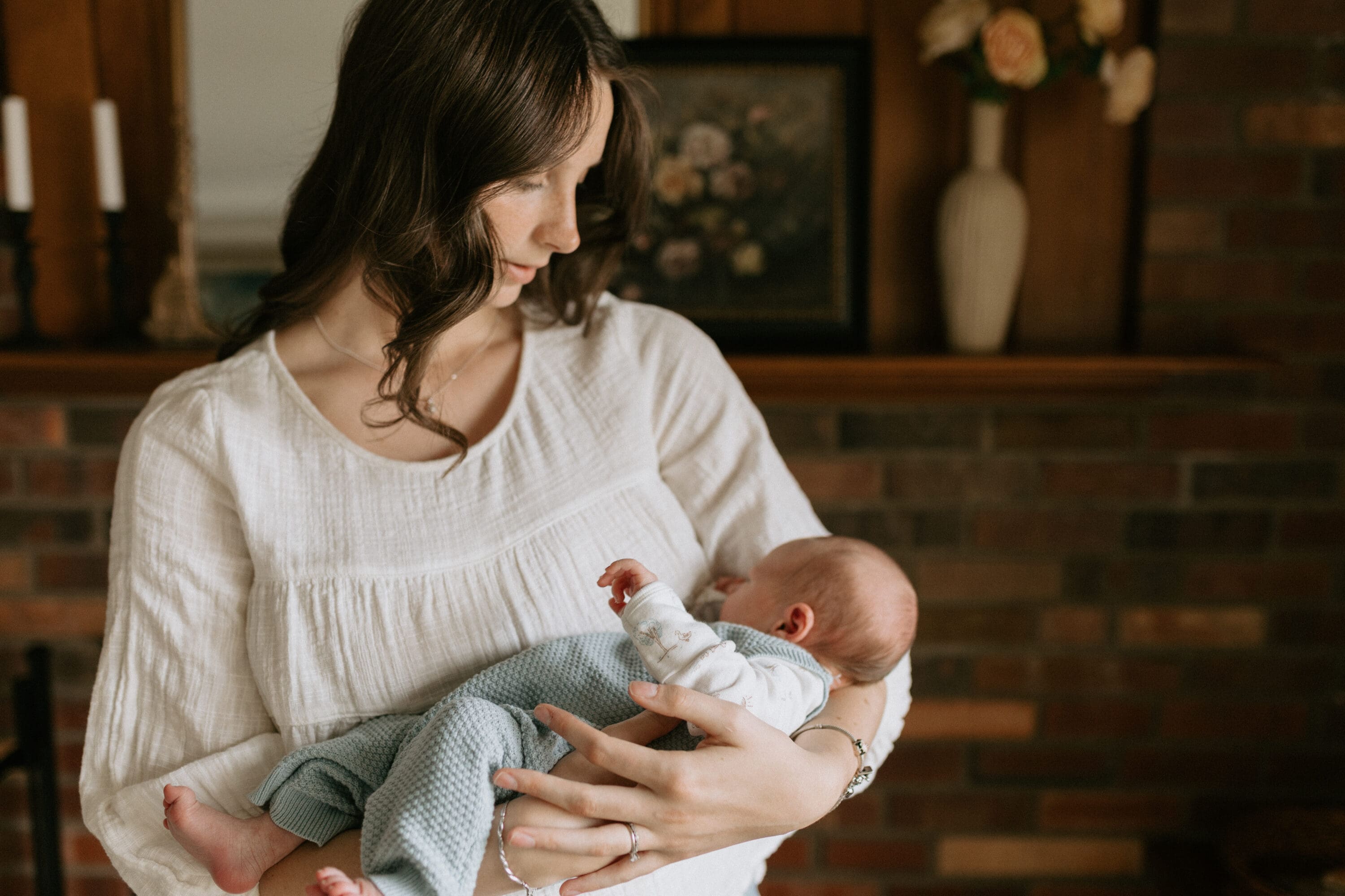 A new mother holds her newborn son. She gazes down at him with a soft expression. She stands in front of their home's brick fireplace decorated with antique and vintage decor.