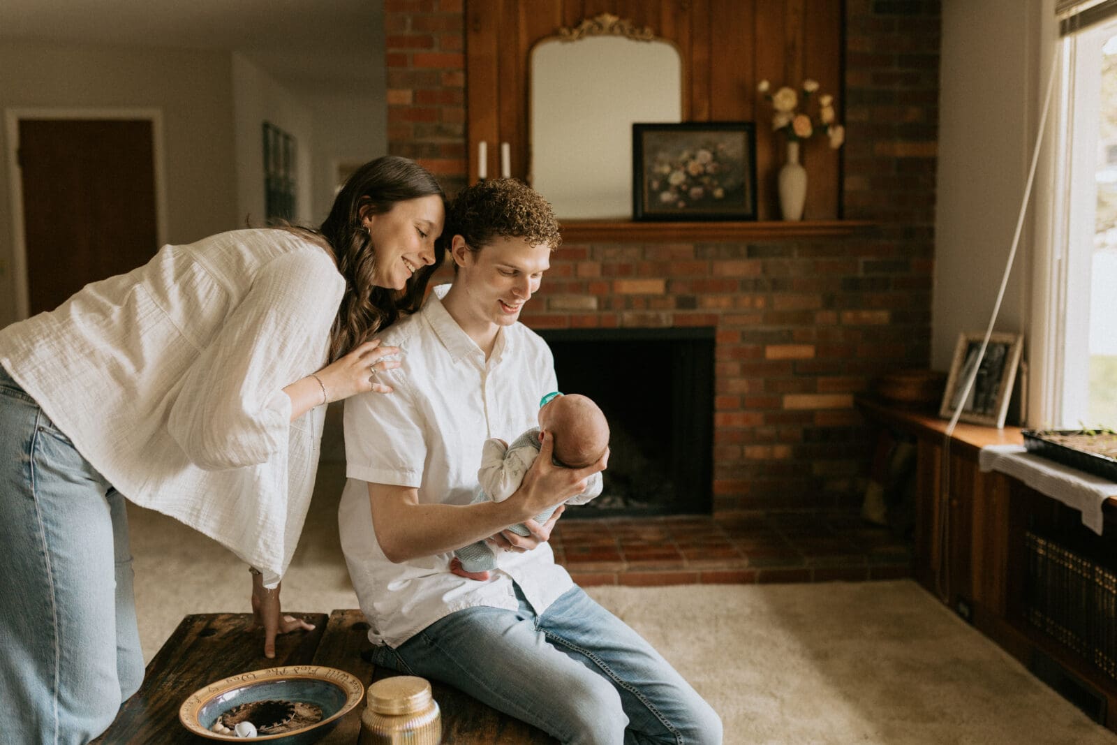 A dad sits on a coffee table looking down at his newborn son. The mom leans over his shoulder. They both smile warmly at the baby, with expressions of amusement.