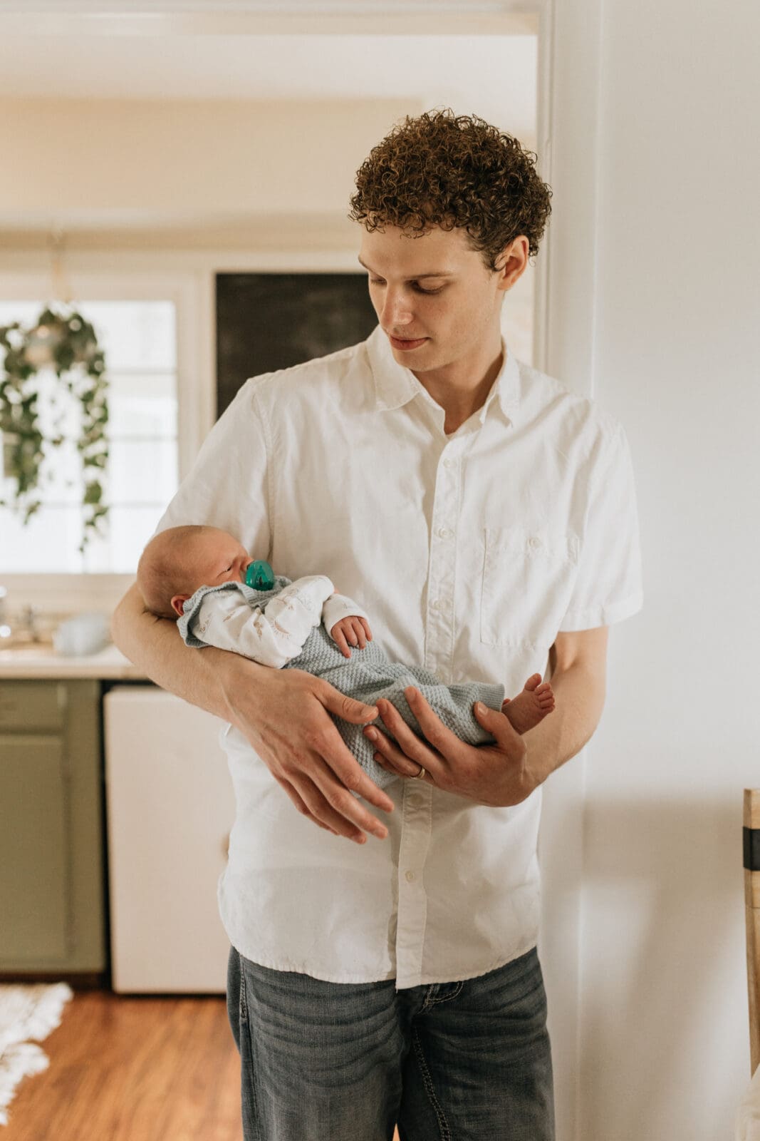 A dad stands in a white doorframe holding his newborn son. He looks down at the baby.