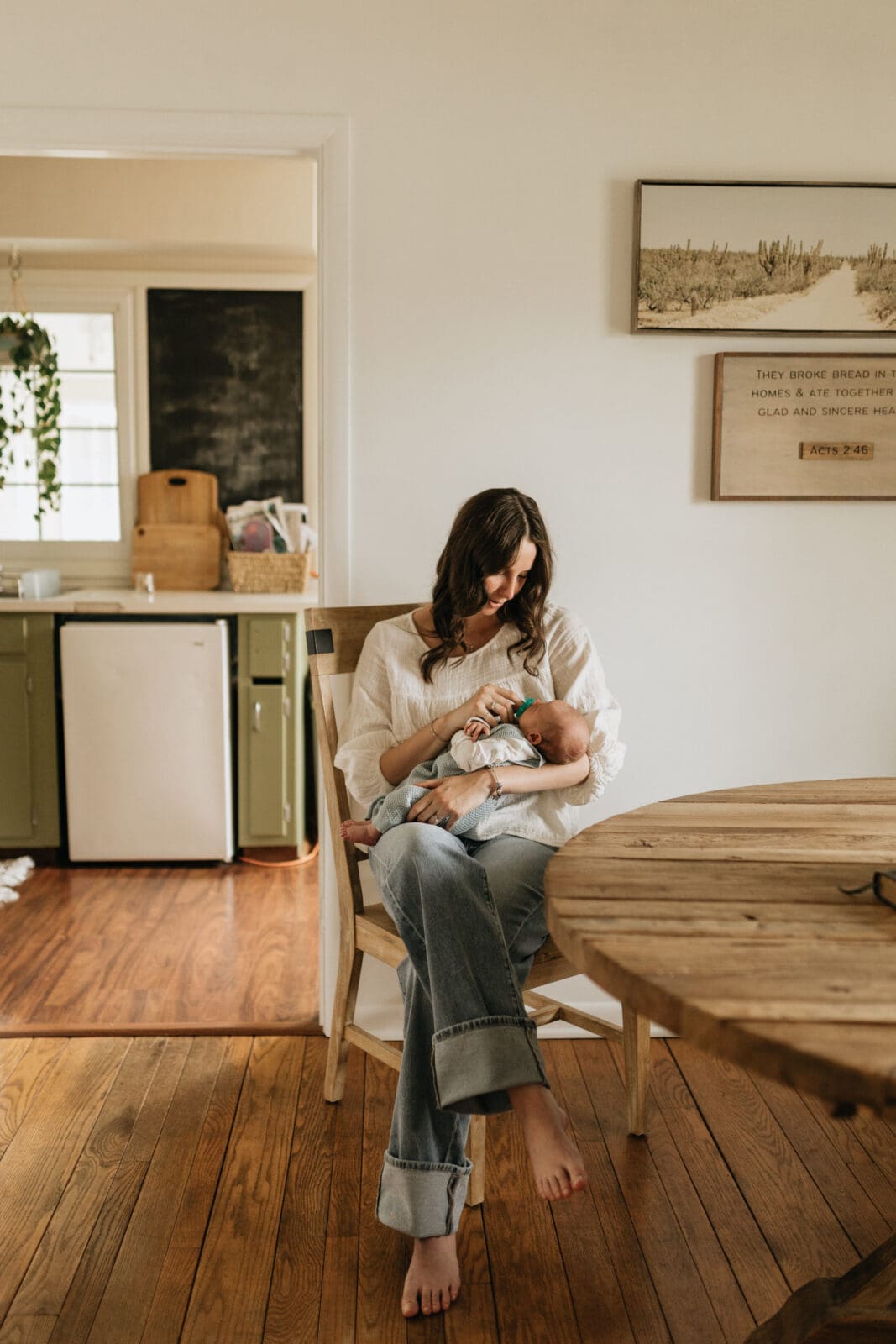 A mom sits in a chair and looks down and her newborn baby son. She is adjusting the pacifier in his mouth to soothe him.