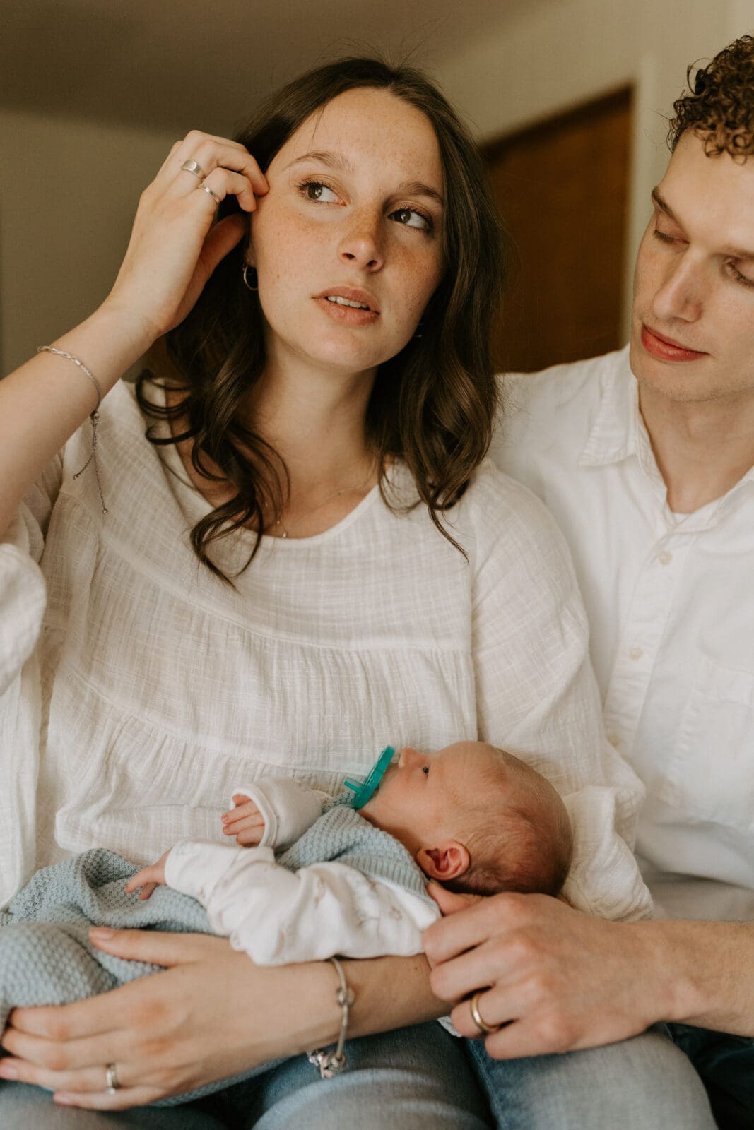 A mother holds her newborn son as she looks away from the camera while brushing some hair out of her eyes. Her husband is looking down at the baby she is holding.