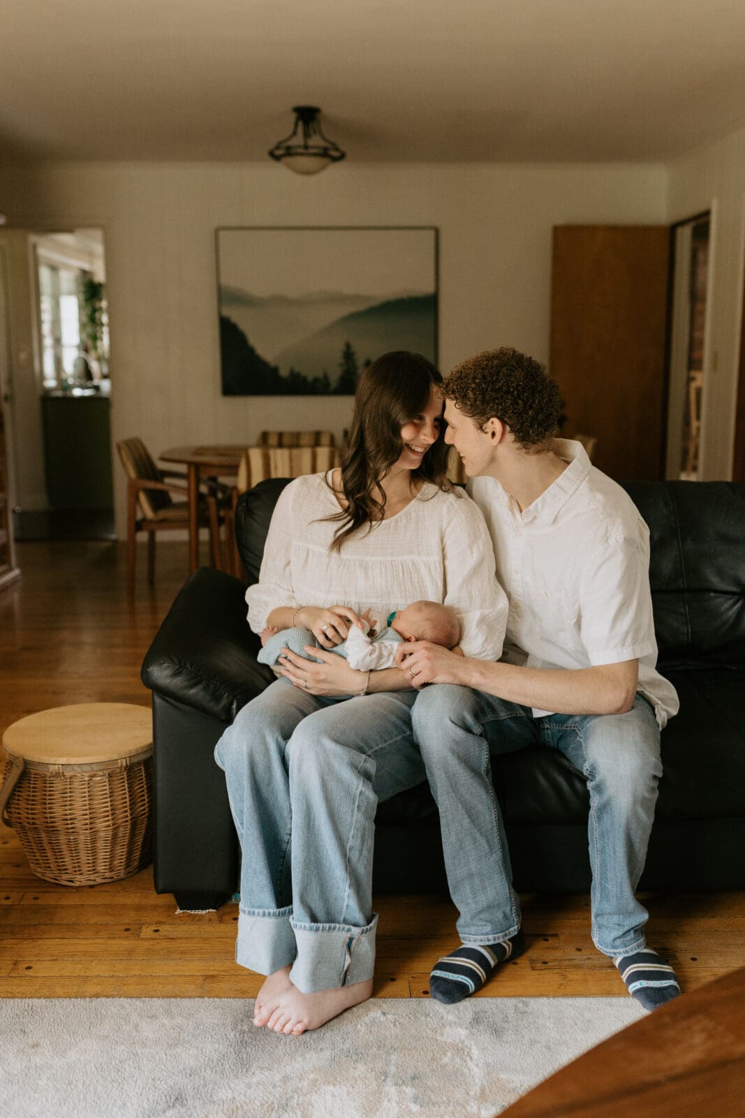 A new mom and dad sit on their living room couch. The mom holds the baby and the new parents put their faces close together and smile at each other.