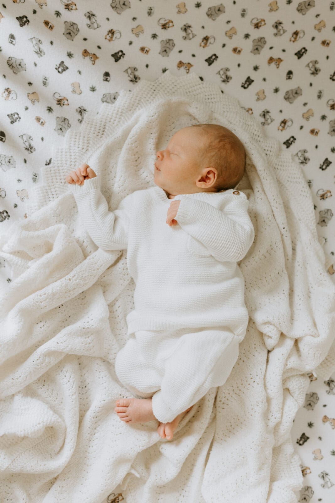 A newborn baby lays in his crib. The sheet has a design with small trucks. The baby is sleeping, dressed in a white knit jumping, laying on a white knit blanket.