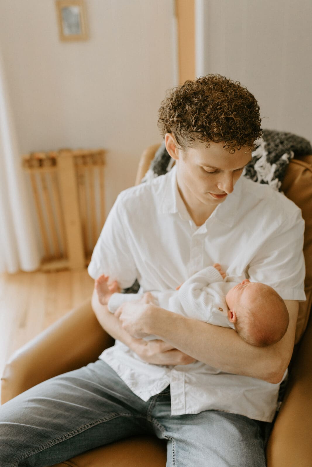 A dad and his infant son are both dressed in white. The dad sits in a chair holding his newborn son, looking at him.