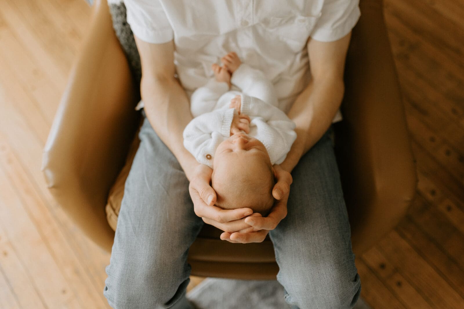 A dad sits in a modern, leather chair holding his newborn baby in his lap. The perspective is from above.