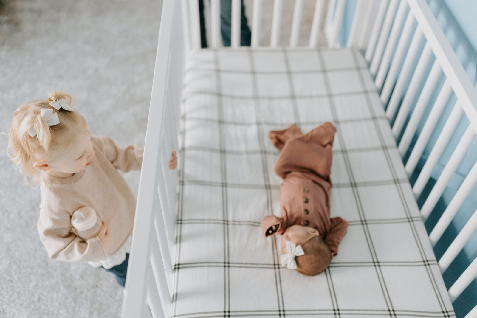 A toddler looks at her infant baby sister sleeping in a crib during a newborn photo shoot with Vanessa Stoller Photography.
