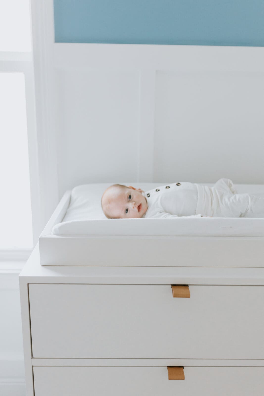 A newborn in a white outfit lays on a white changing table looking at the camera. The room is light blue and white and the furniture is modern.