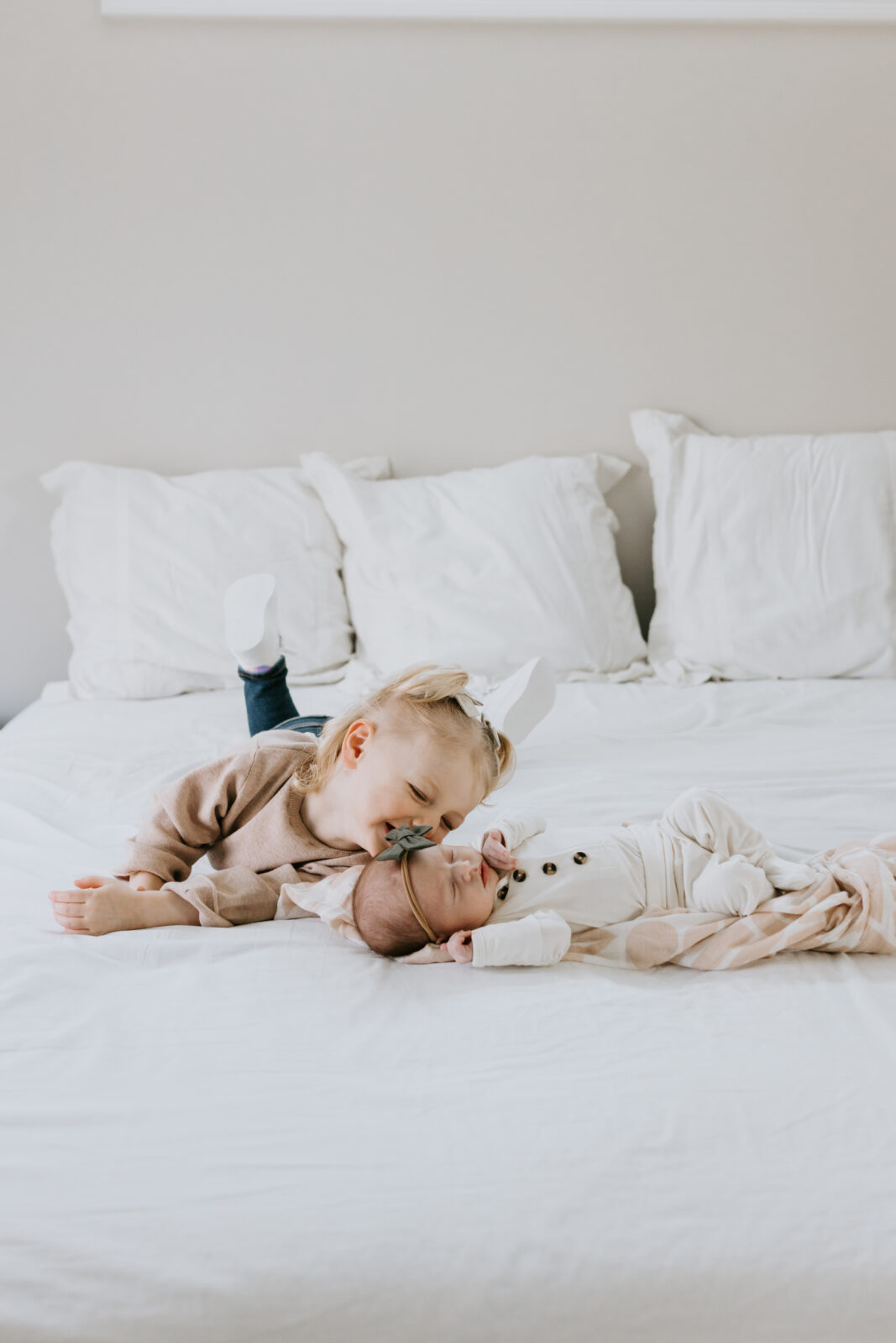 A baby photo session where a toddler lays on the bed next to her newborn sister and playfully smiles at her sleeping baby sister.
