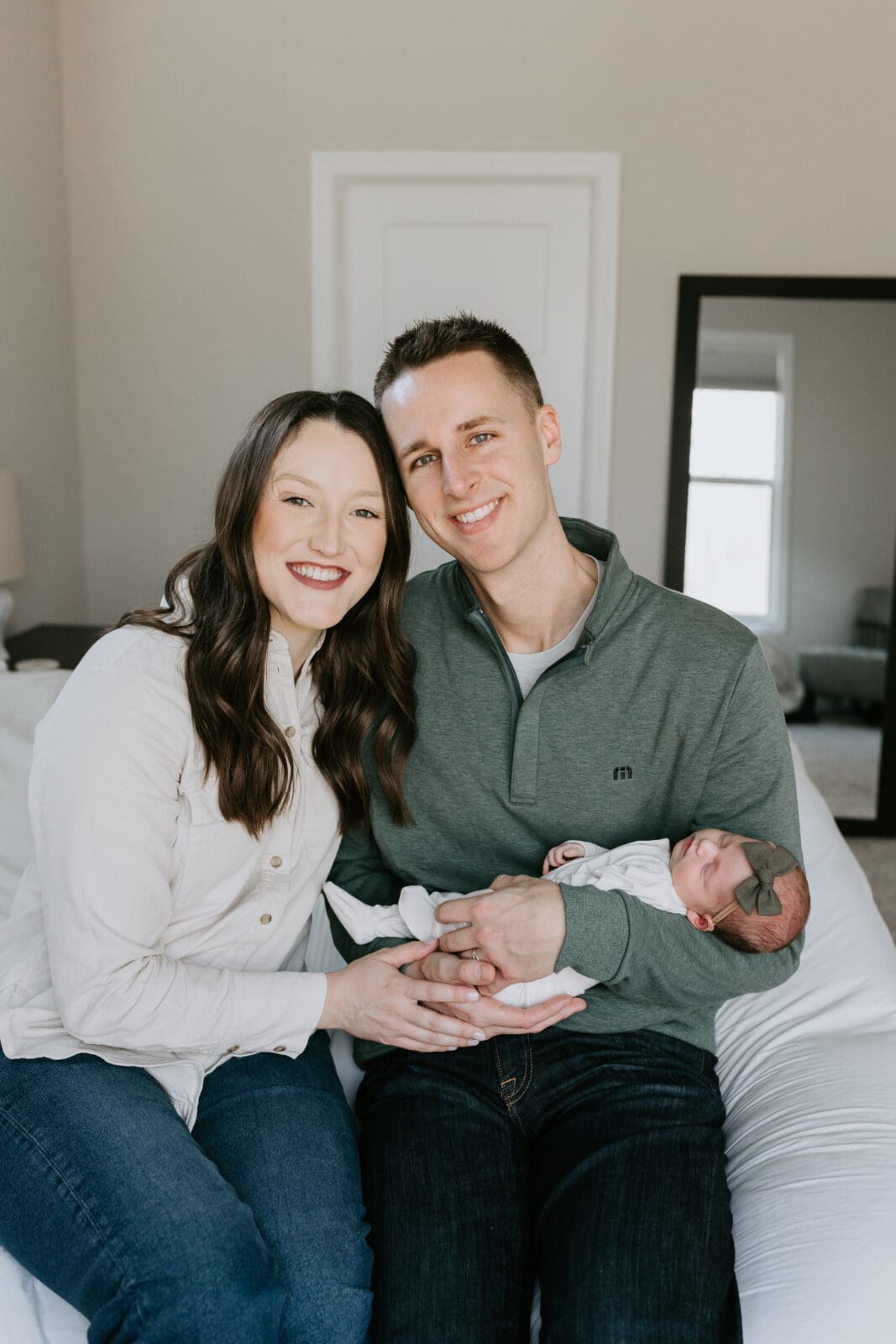 A mom and dad smile and look at the camera while holding their new sleeping newborn daughter. The sit on the edge of their bed at their West Lafayette, Indiana home for their newborn photo shoot.