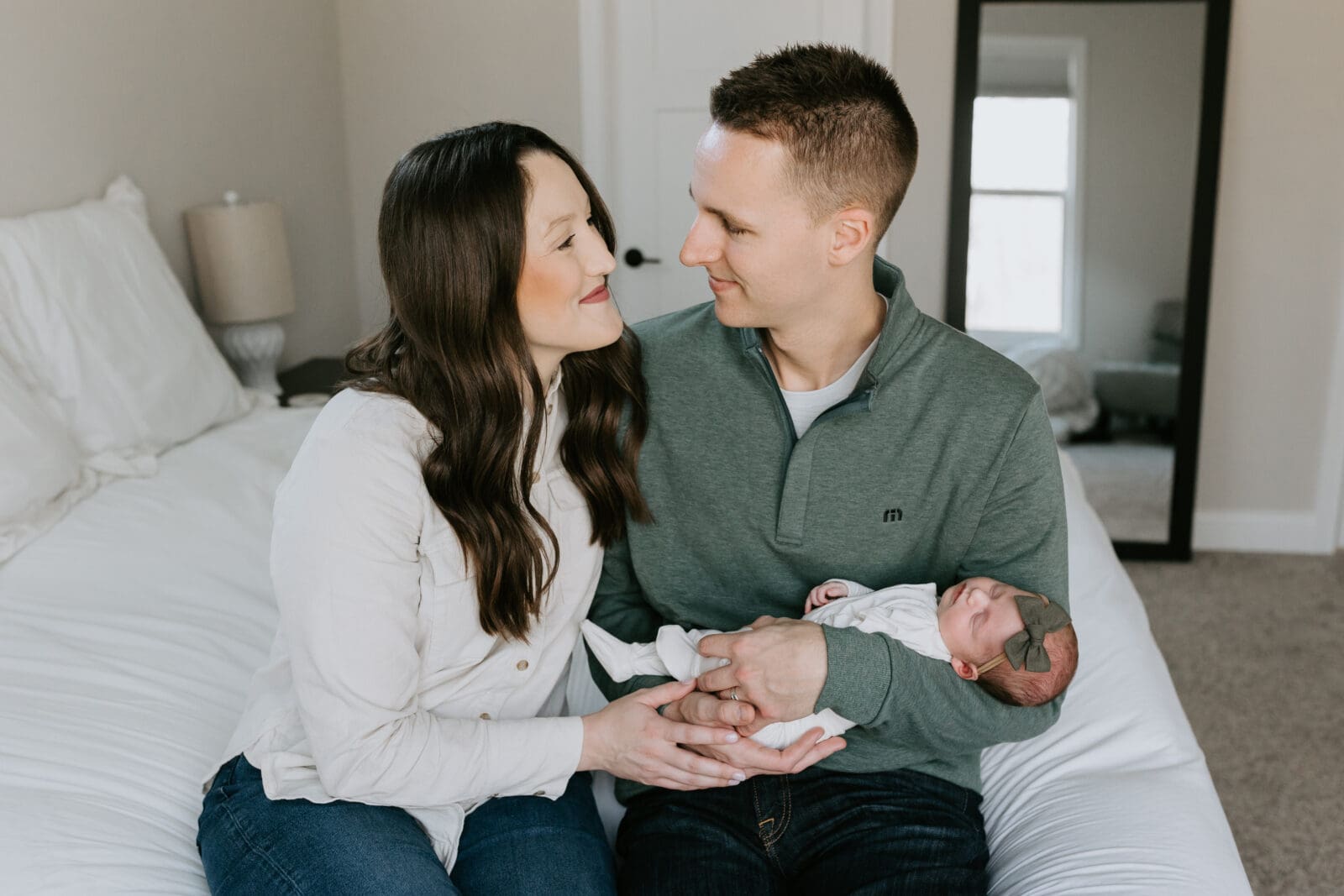 A dad and mom look at each other and smile while the dad holds their sleeping infant daughter. This is taken in their bedroom for their lifestyle newborn photo session with Vanessa Stoller Photography.