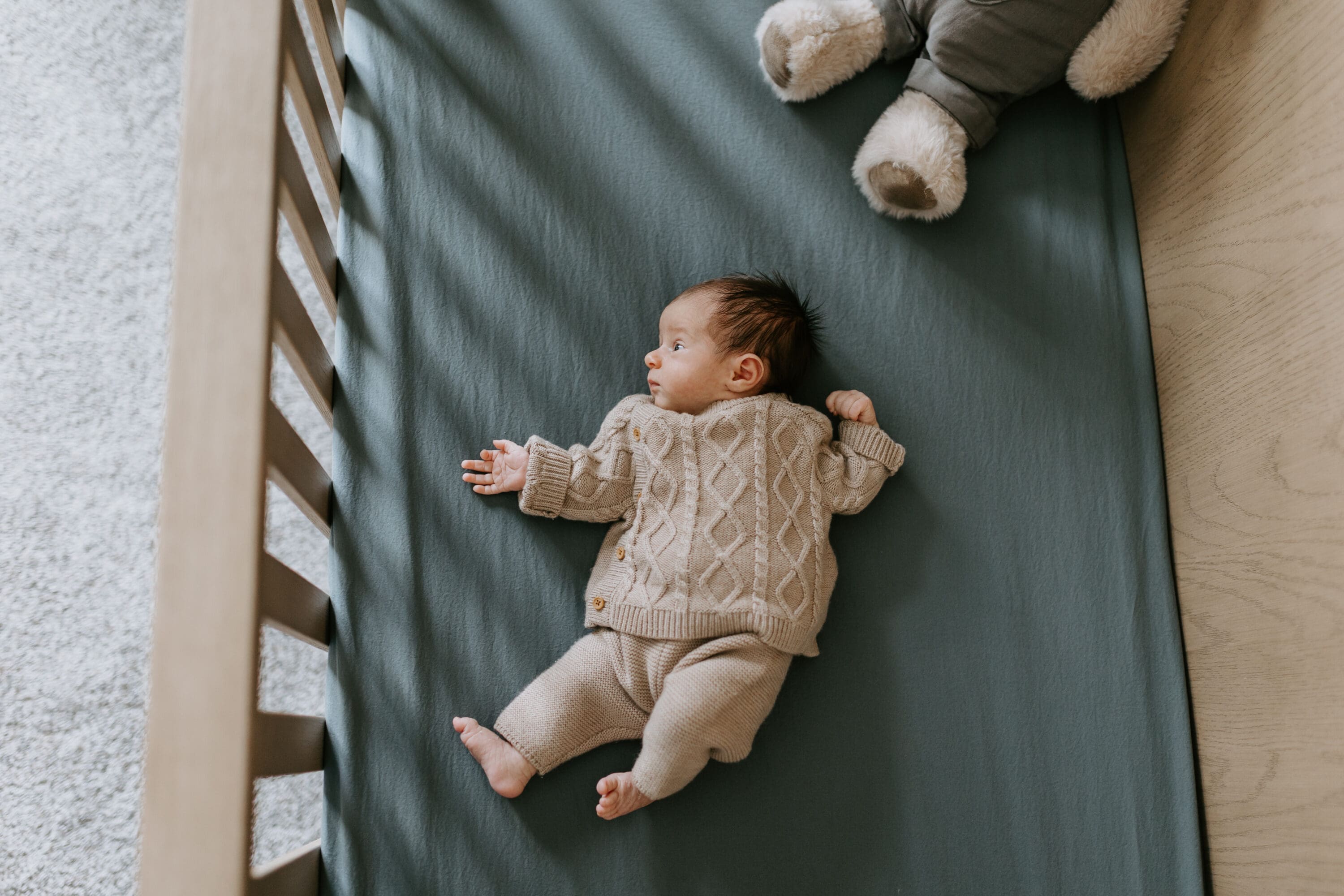 A newborn baby boy lays in his crib. The sheets are a soft blue and a large stuffed animal can be seen in the corner of the frame. The baby is awake but content in for baby pictures in lafayette, indiana by Vanessa Stoller Photography.