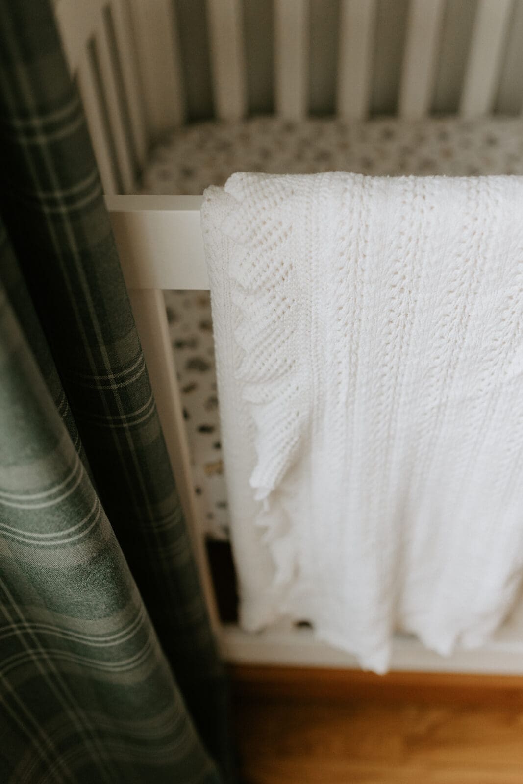 A white heirloom knitted baby blanket hangs folded over the side of a white crib. The dark green curtains on the window next to the crib is also in the frame.