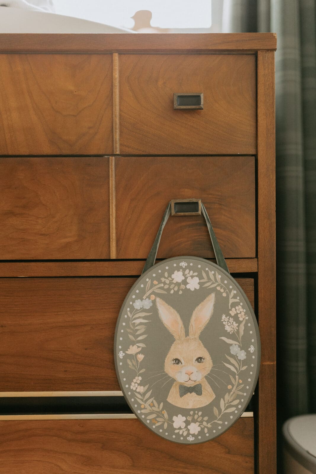 A wooden plaque with a painting of a bunny with a green velvet ribbon hangs from the knob of a vintage dresser in a baby nursery.