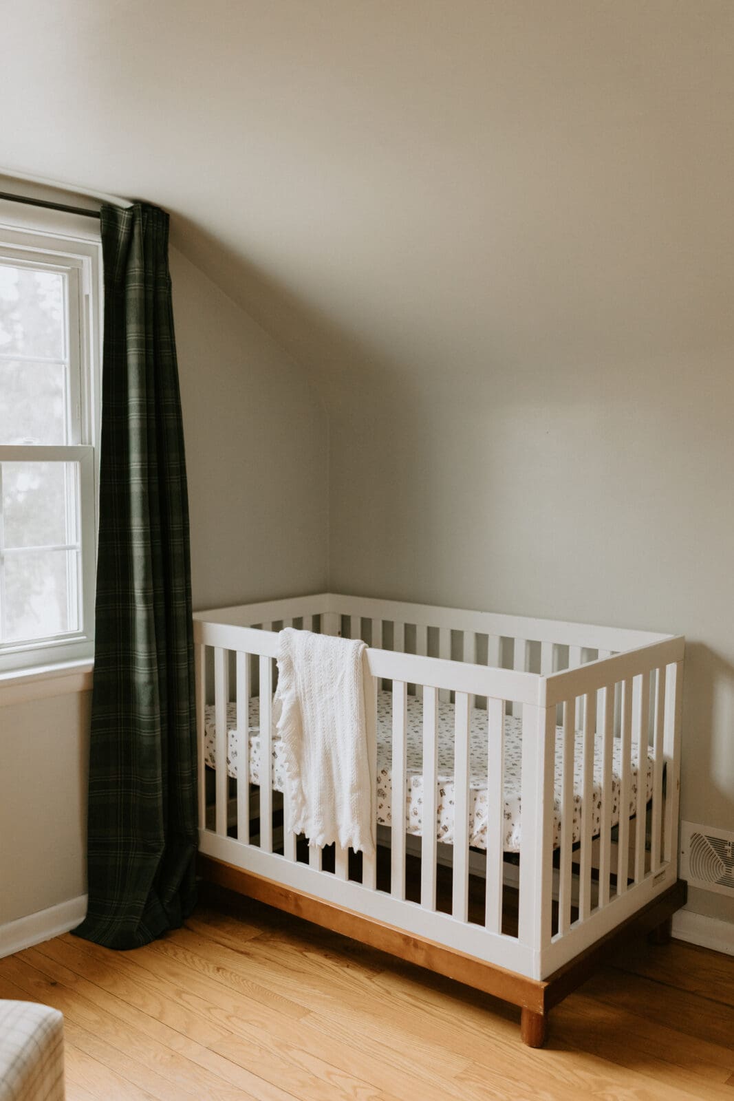 A crib in the corner of the nursery with natural window light. The crib is white and has a knitted blanket folded and hanging over the side.