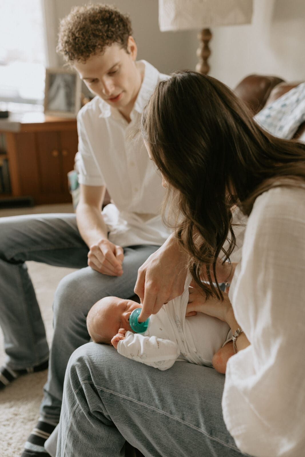 A lifestyle newborn photography moment where a mom and dad sit together on a couch trying to calm their newborn baby with a pacifier.