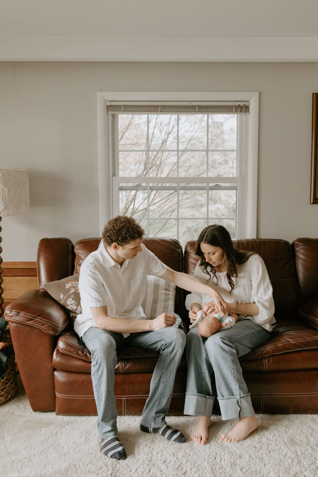 A new mom and dad sit on their couch soothing their newborn baby. This is a lifestyle photo session in Lafayette, Indiana by Vanessa Stoller Photogaphy.