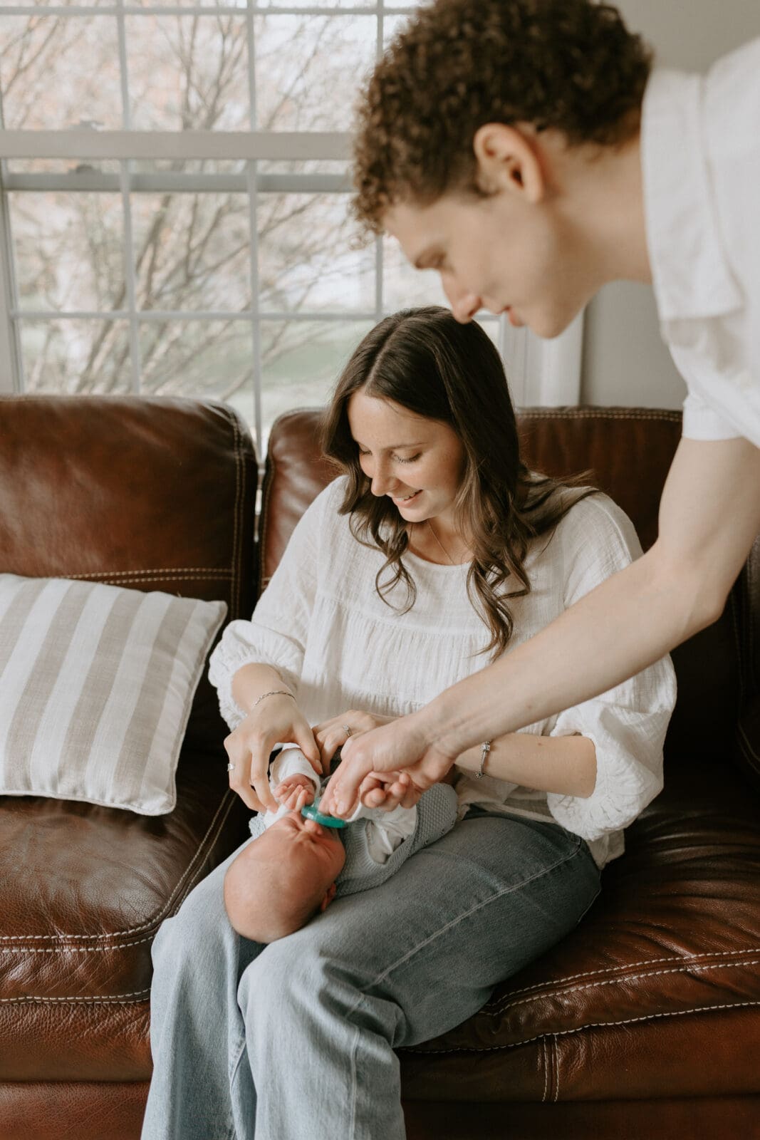 A mom holds a baby on her lap while the dad tries to soothe the baby with a pacifier. The mood is lighthearted and the parents are smiling.
