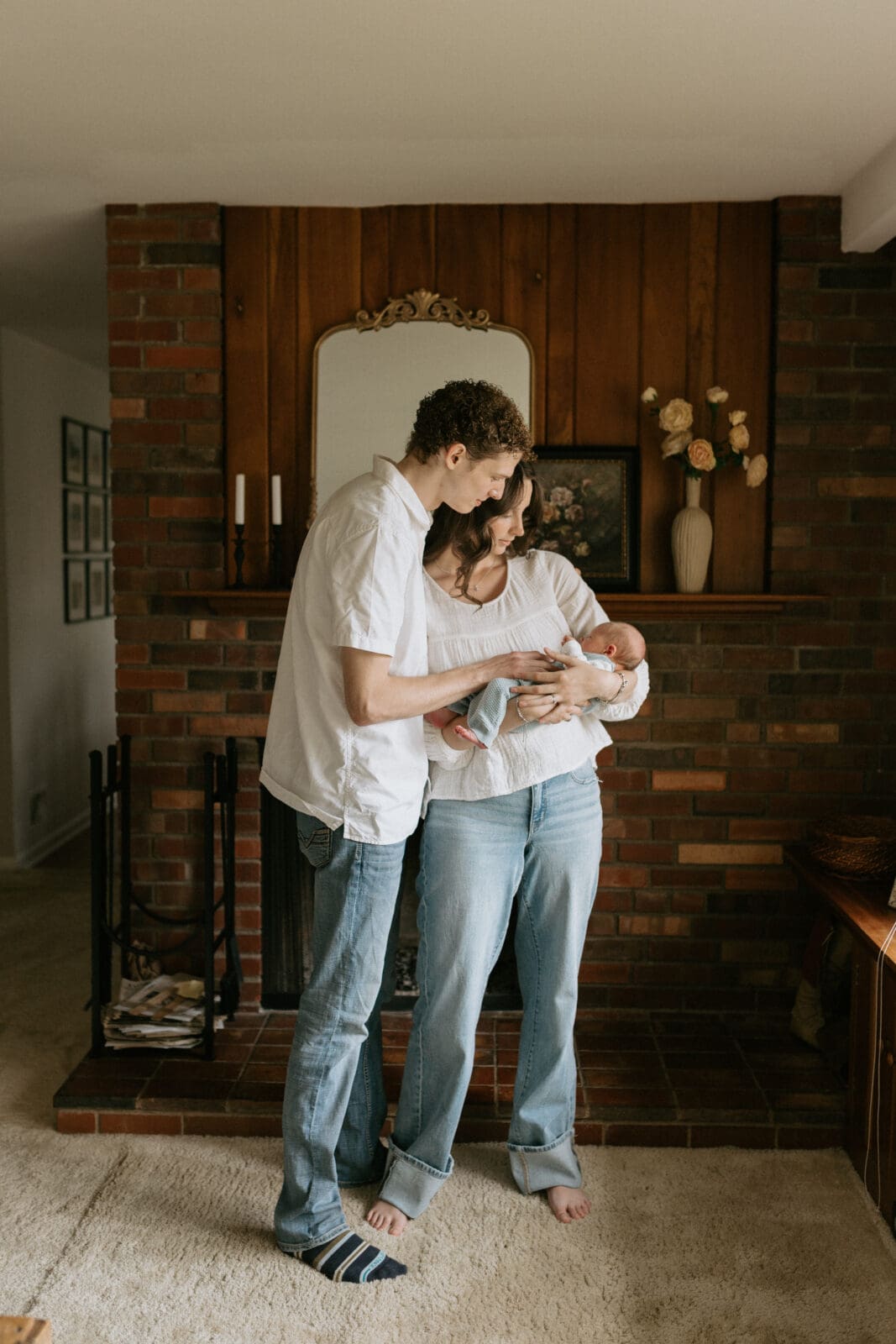 Parents stand in window light in front of their fireplace gazing down at their newborn baby.