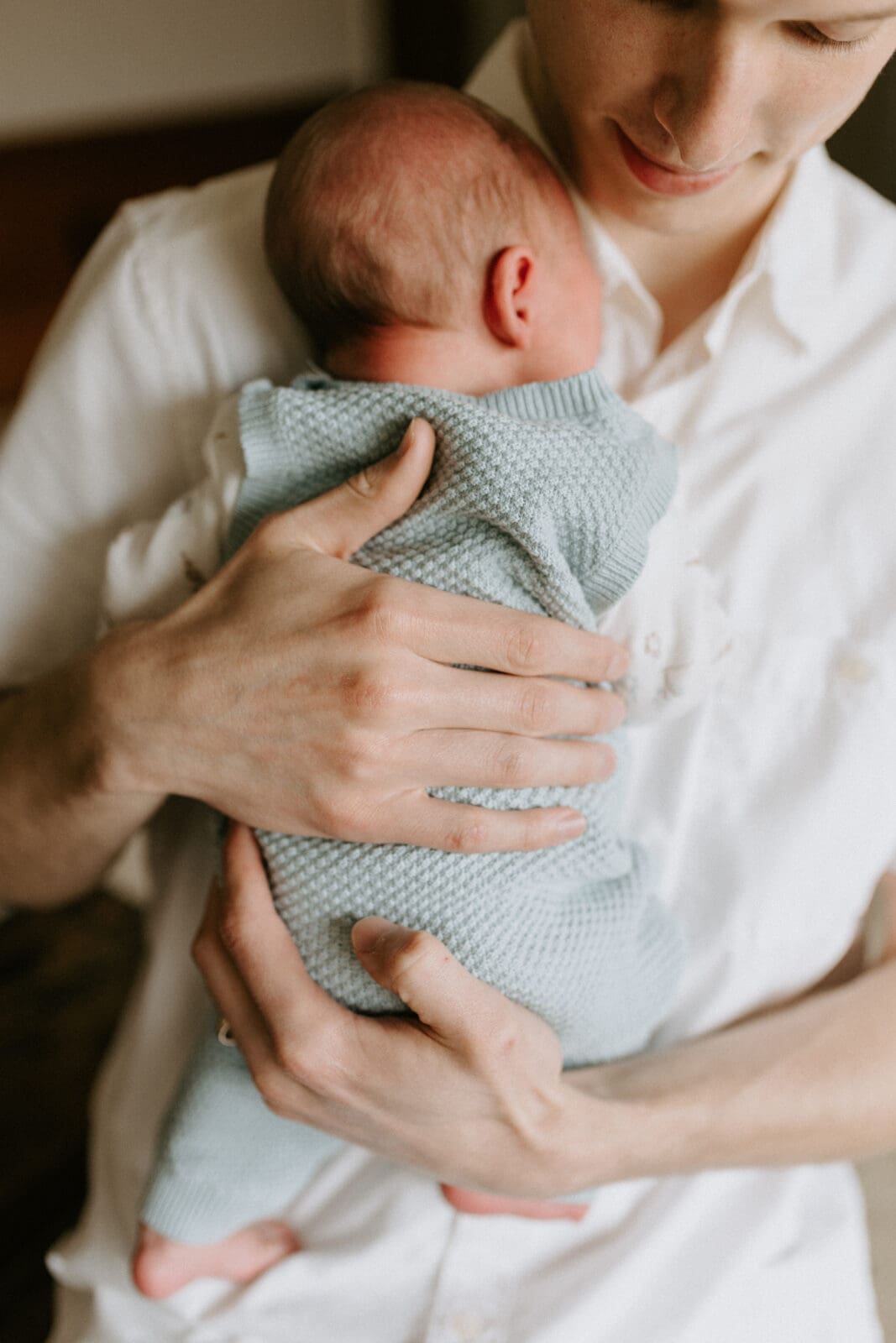 A new dad hold his infant son close. The baby wears a blue knit jumper and the dad wears a white button up shirt.
