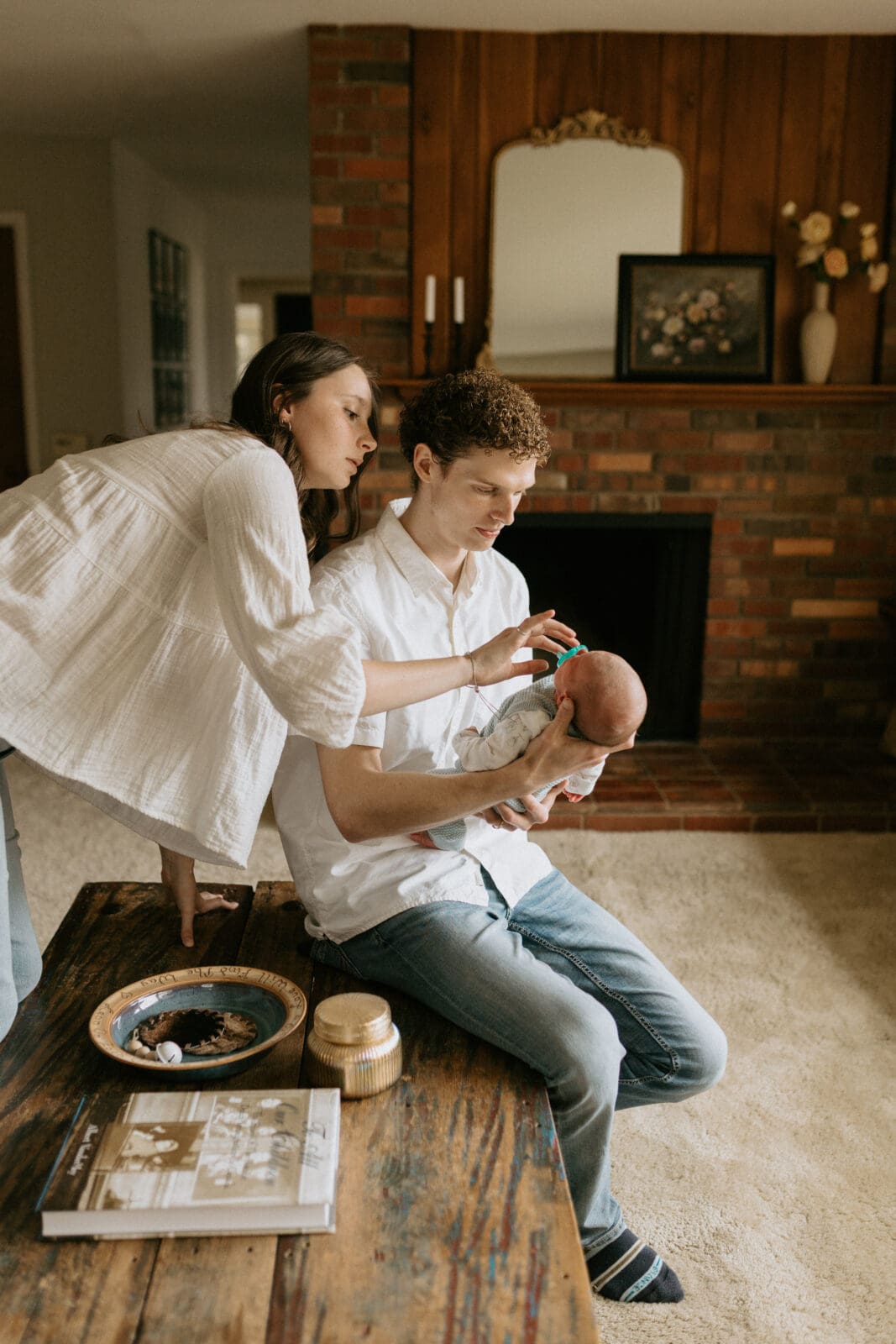 A dad sits on a coffee table in the window light. He holds his newborn son in his arms while the mom leans over the dads shoulder, attempting to soothe the baby with a pacifier.