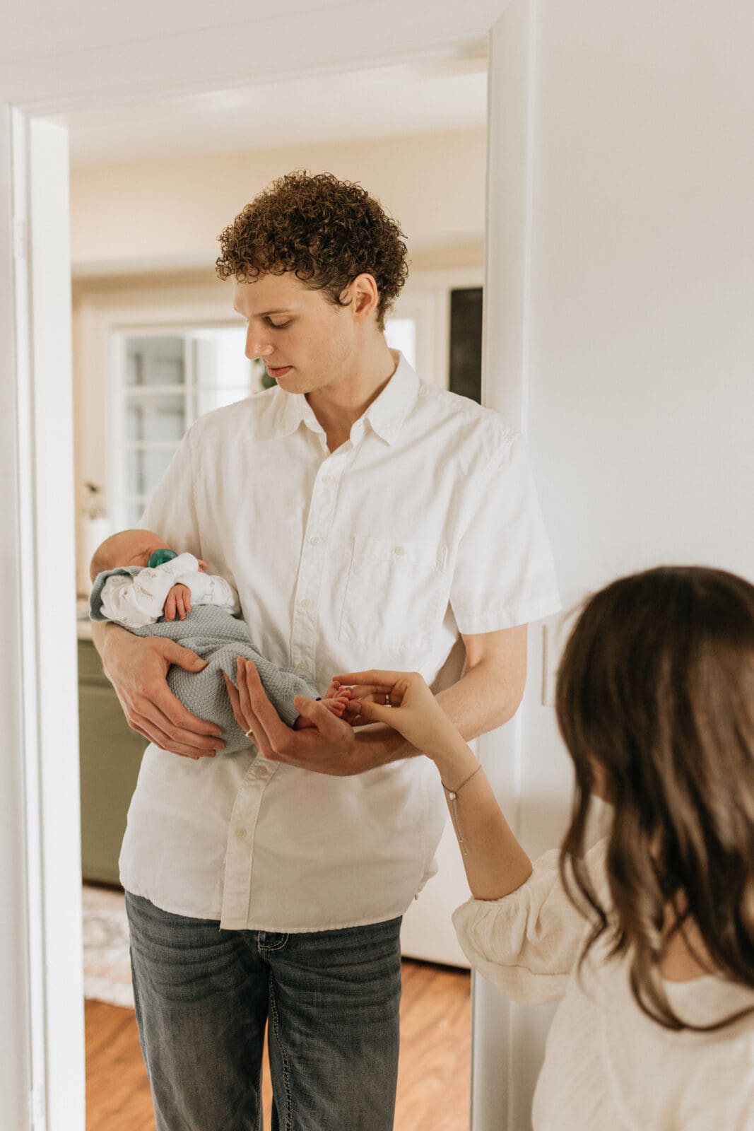 A dad stands in a doorframe looking down at the newborn son he is holding. The mom is in the corner of the frame, reaching to touch the baby's toes.