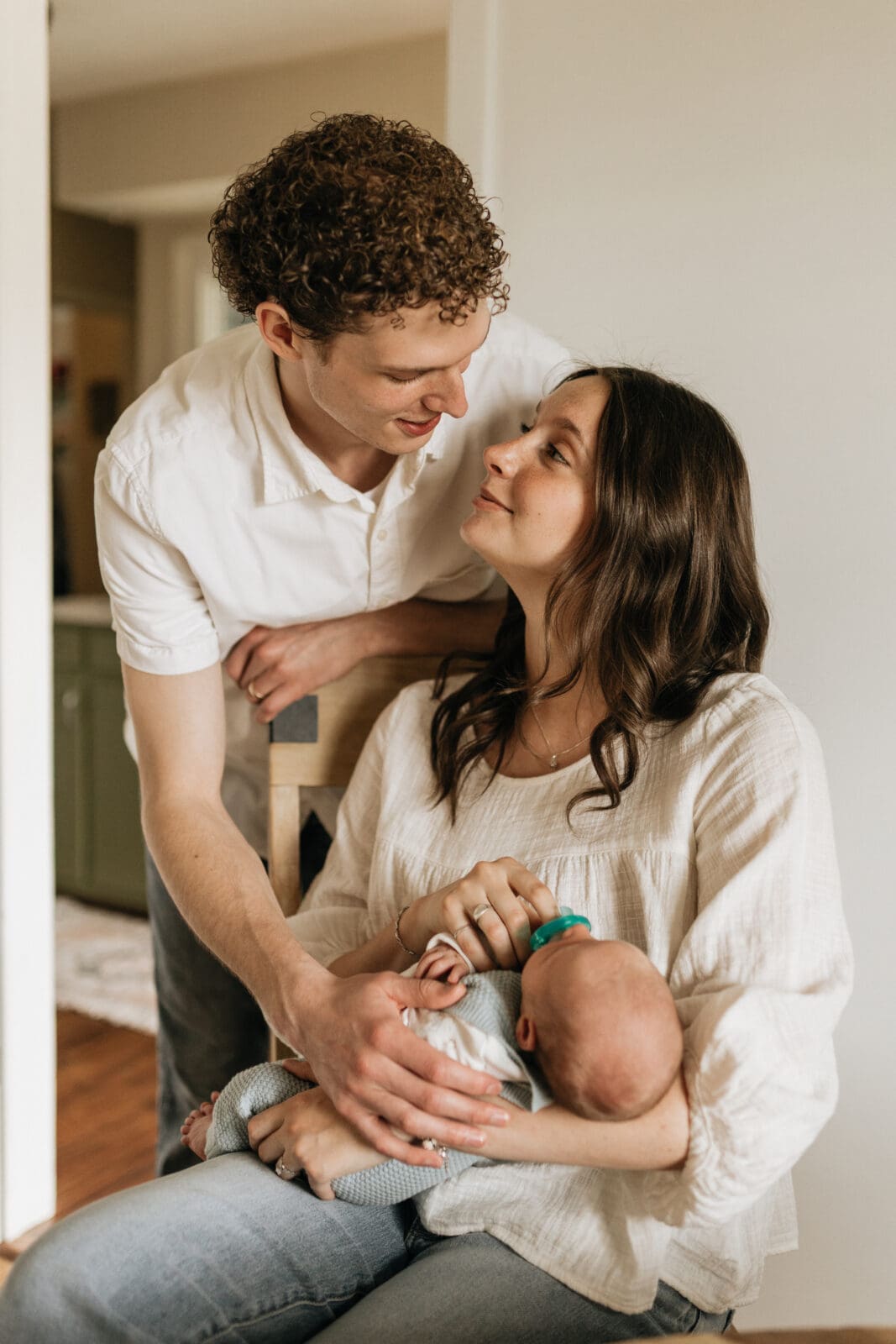 A mom and dad look lovingly at each other while holding their newborn baby in their arms.