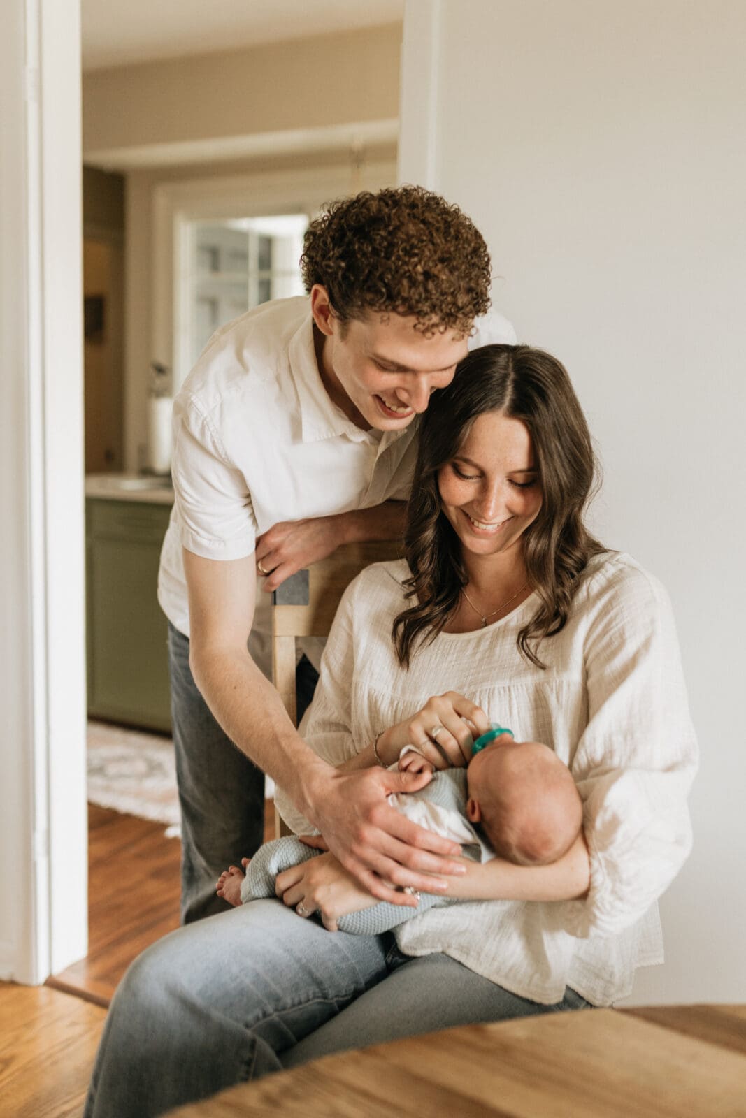 A new mom and new dad come together at the dining room table and look down at their newborn baby with smiles on their faces.