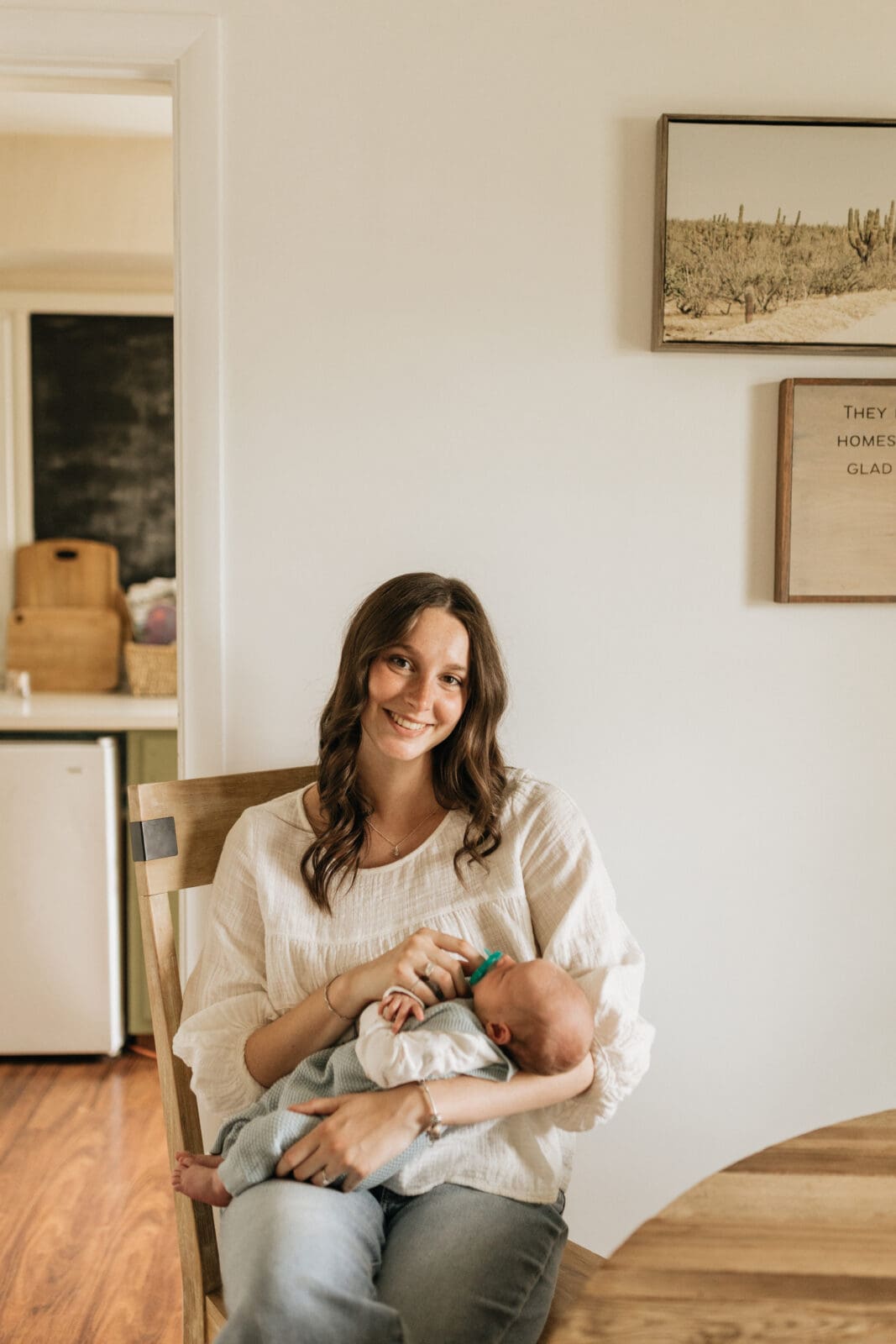A new mom sits at her dining room table. She is holding her infant son and smiling and looking at the camera for newborn photos with Vanessa Stoller Photography.