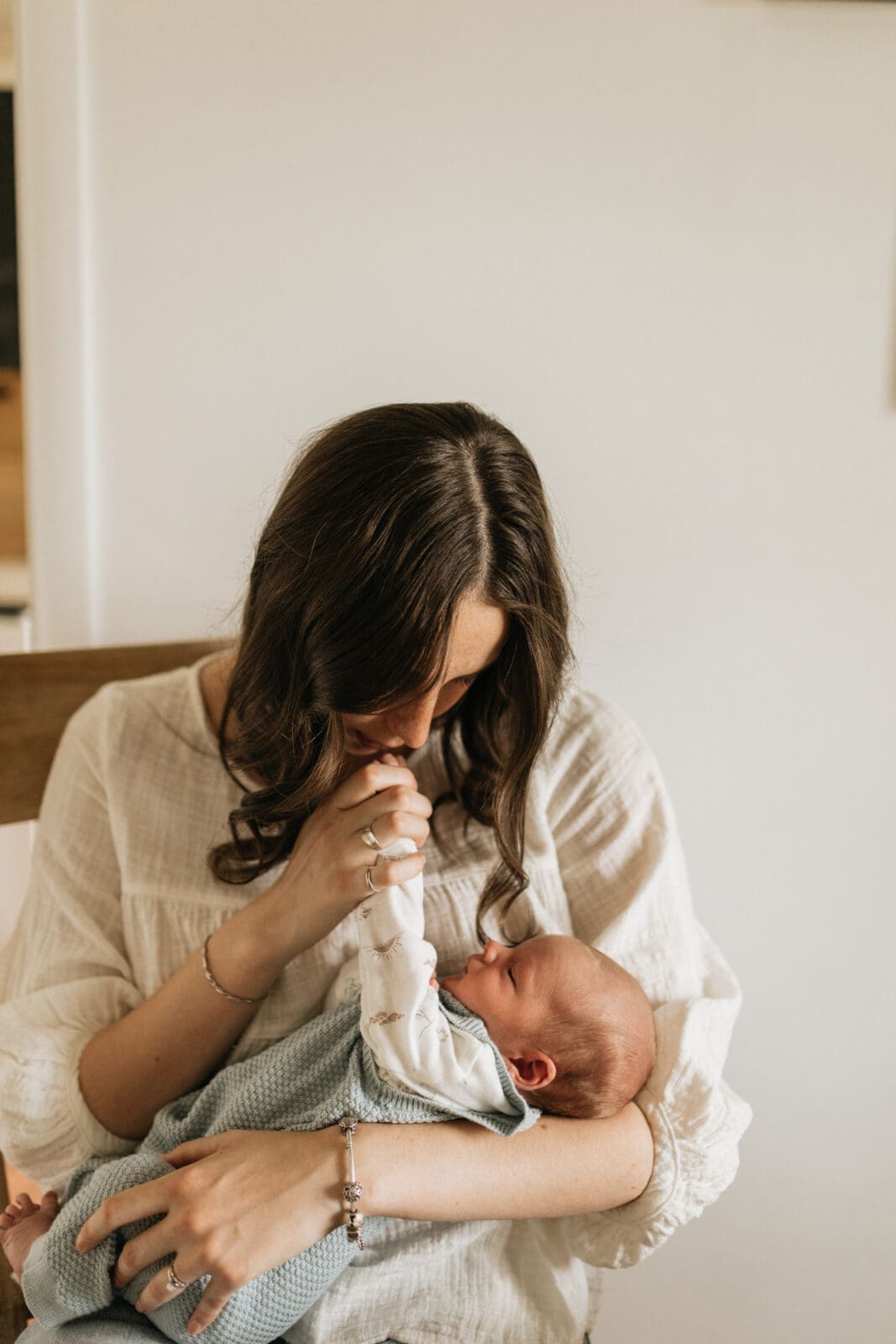 A mom sits in a chair holding a newborn baby. She is lifting his hand to her lips and giving his fingers a kiss.