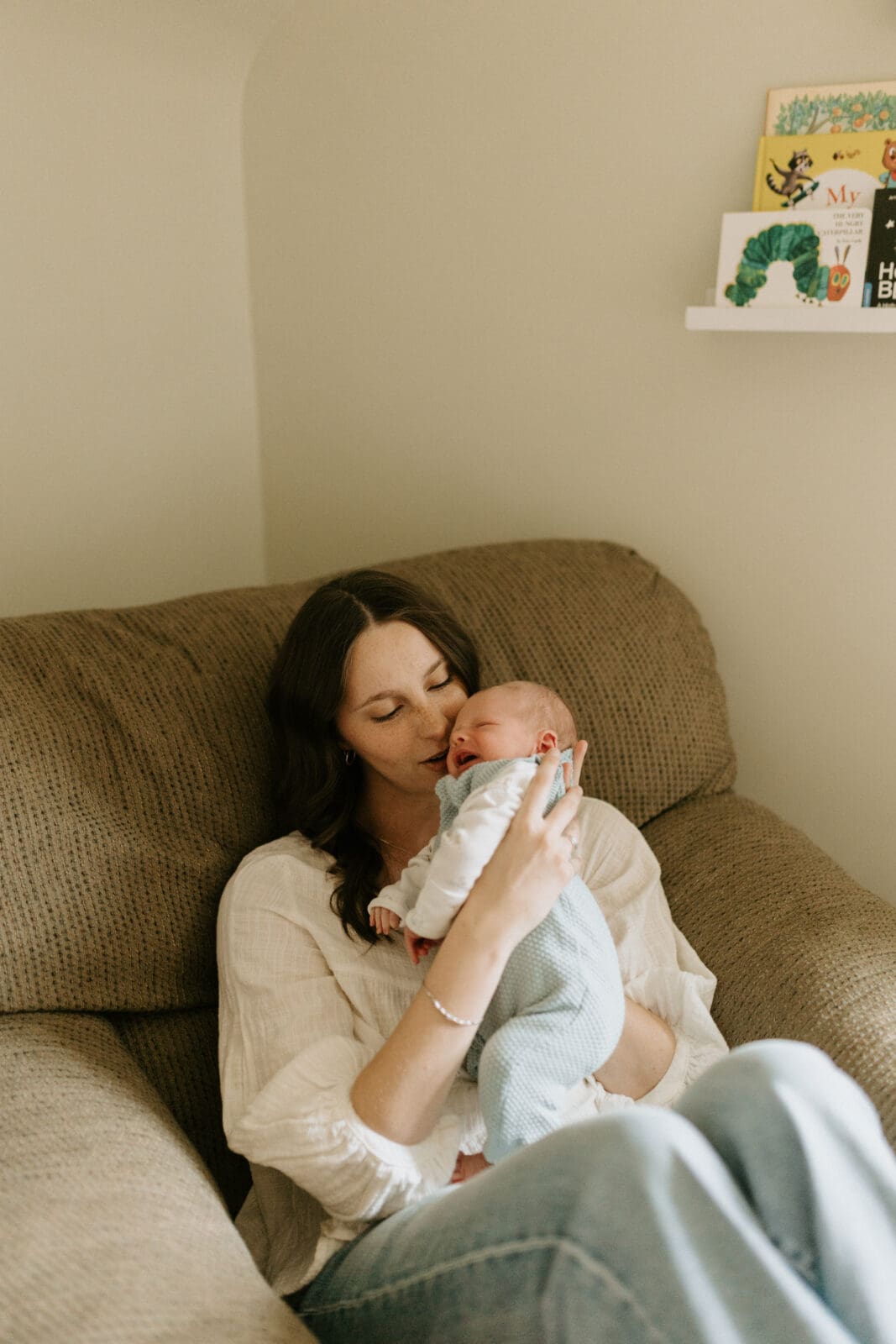 A mom sits in a large chair in the baby's nursery. She is trying to soothe the crying baby and is smiling slightly.