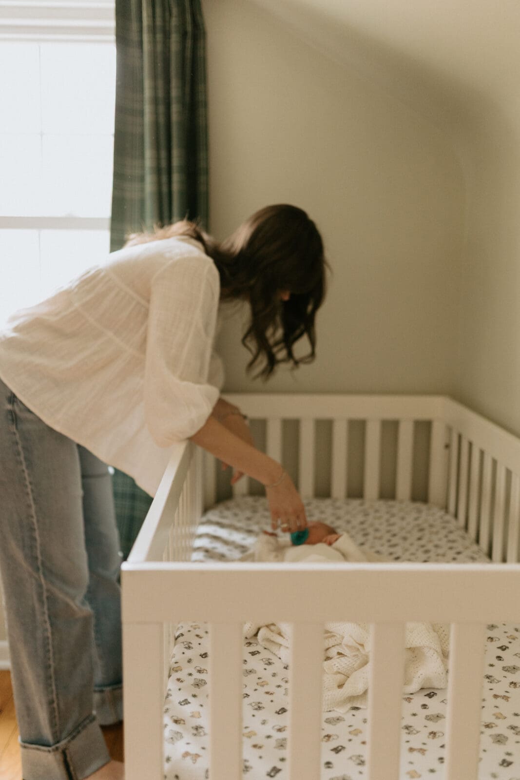 A mother leans over the side of a baby's crib. She is trying to sooth her newborn with a pacifier as he lays in the crib.