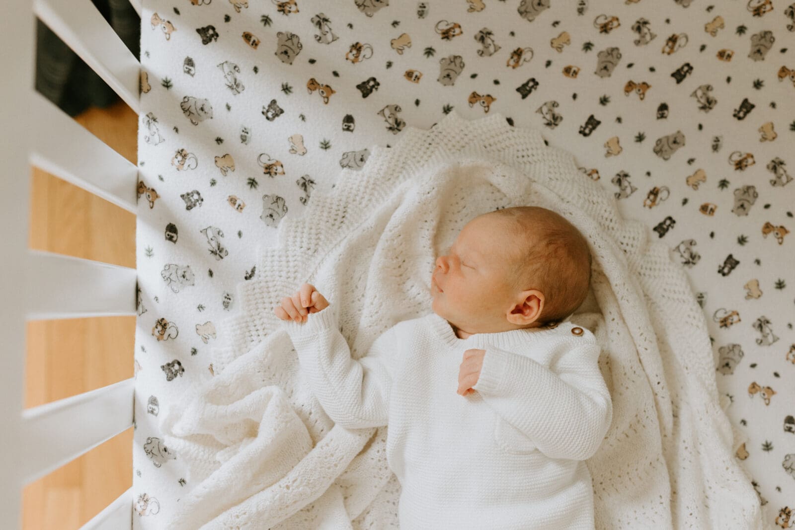 A baby sleeps in his crib. He is dressed in white laying on a white blanket.