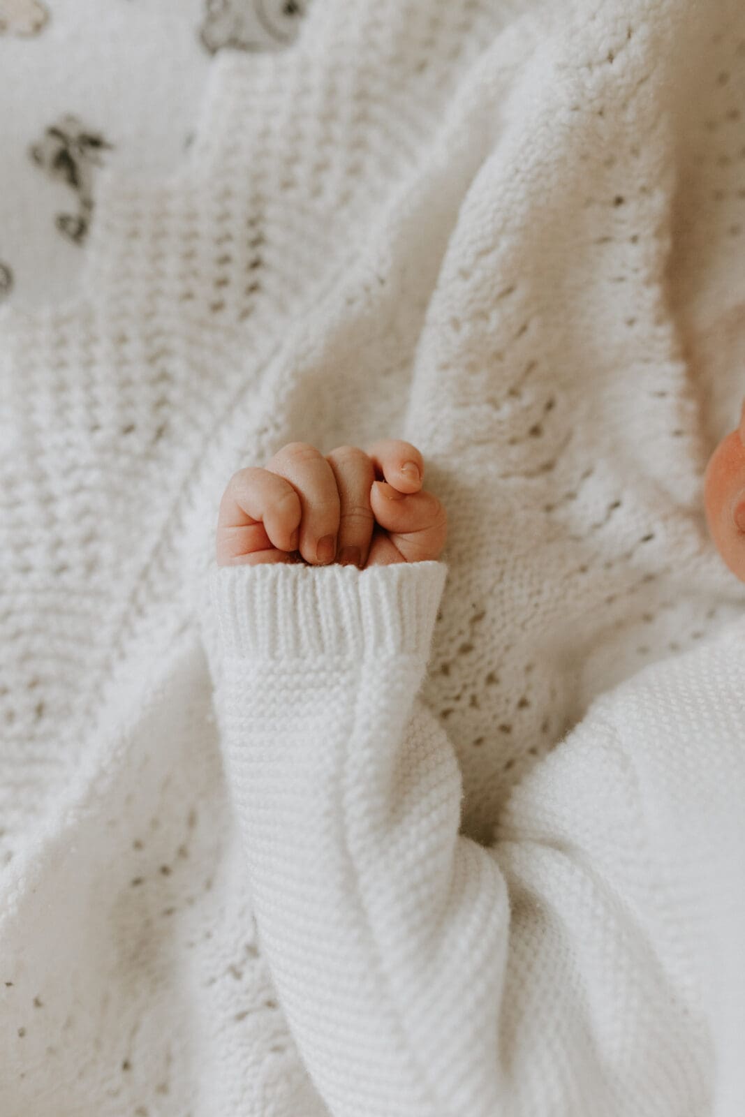 A close up image of a baby's hand and fingers. The baby wears a white jumper and is sleeping on a white knit blanket.