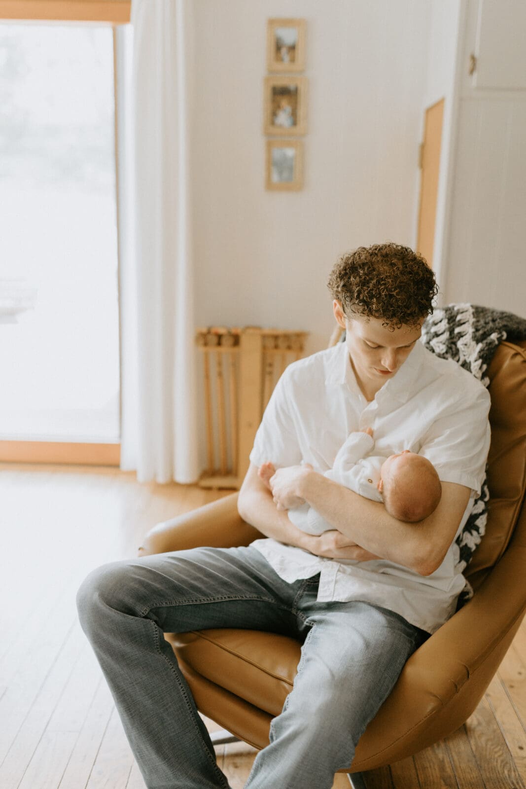 A dad sits in a mid century vintage chair and looks down at the newborn baby boy he is holding.