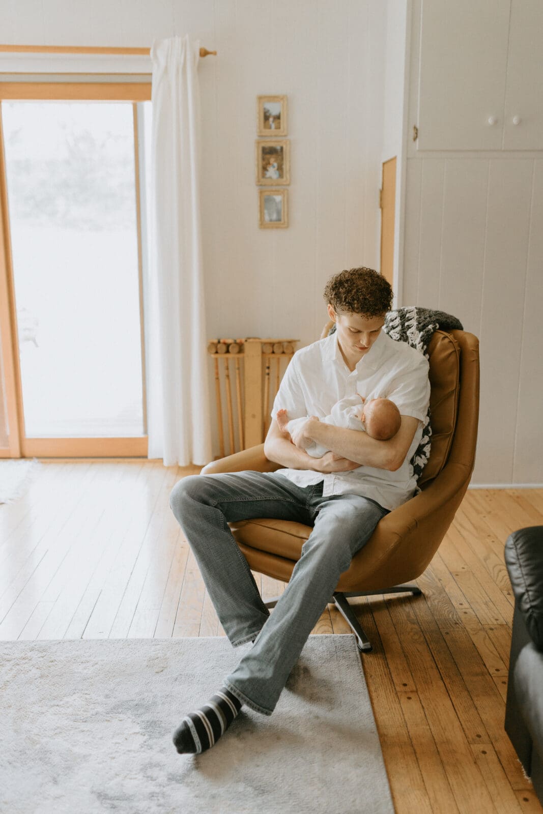 A dad sits in a tan mid century vintage chair and holds his baby boy. The dad has a peaceful look on his face as he gazes down at his baby. Both are dressed in white.