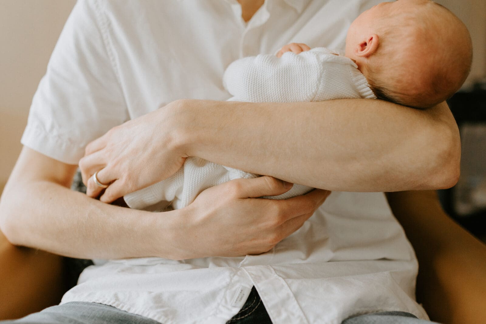 A close up image of a dad cradling his newborn baby son in his arms. Both are wearing white clothing.
