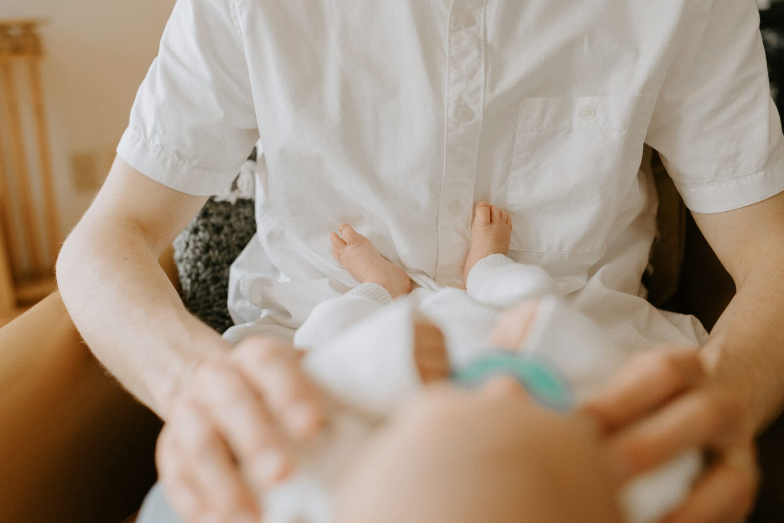 A baby lays on his dad's lap. The camera focuses on the baby's does that rest on his dad's chest.
