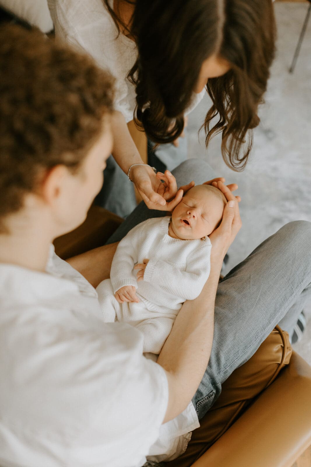 A dad holds his infant son in his lap and looks down at him. The mom stands over the baby and gently touches his cheek with her finger. The camera angle is from above.