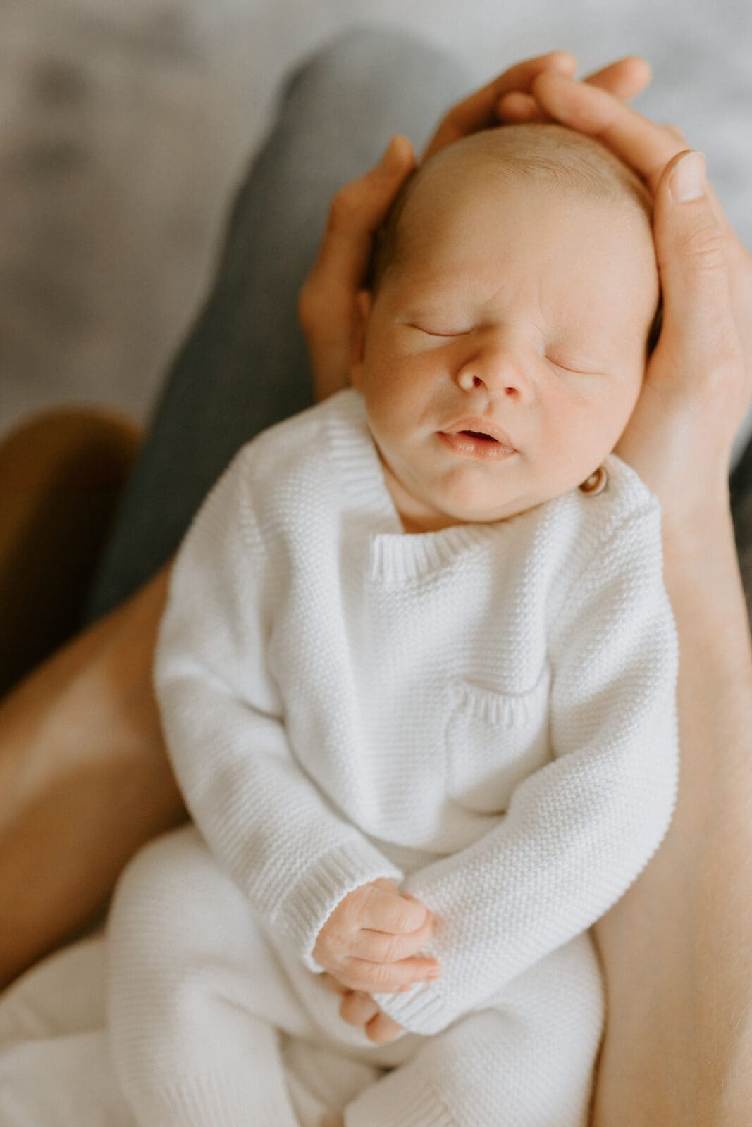 A baby is sleeping in his father's lap. The father's hands cradle the baby's head. The baby wears a white jumper.