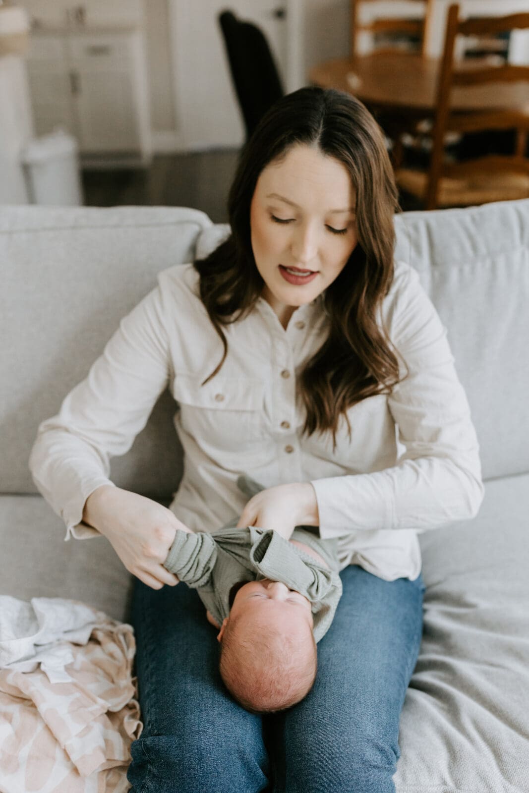 A new mom sits on a sofa and changes her newborn baby's clothes.