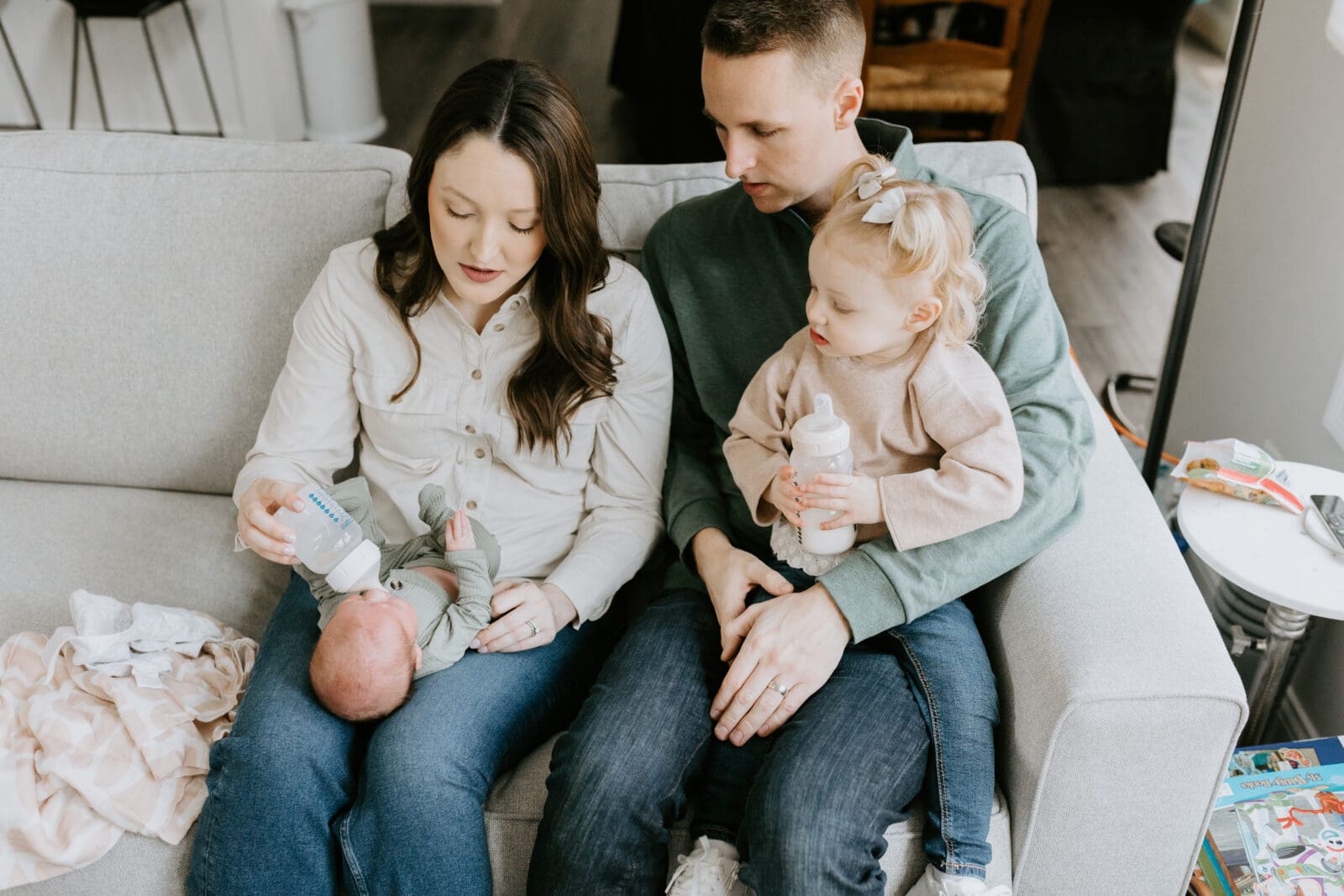 A family of four sits on a sofa while the mom feeds the newborn baby a bottle.