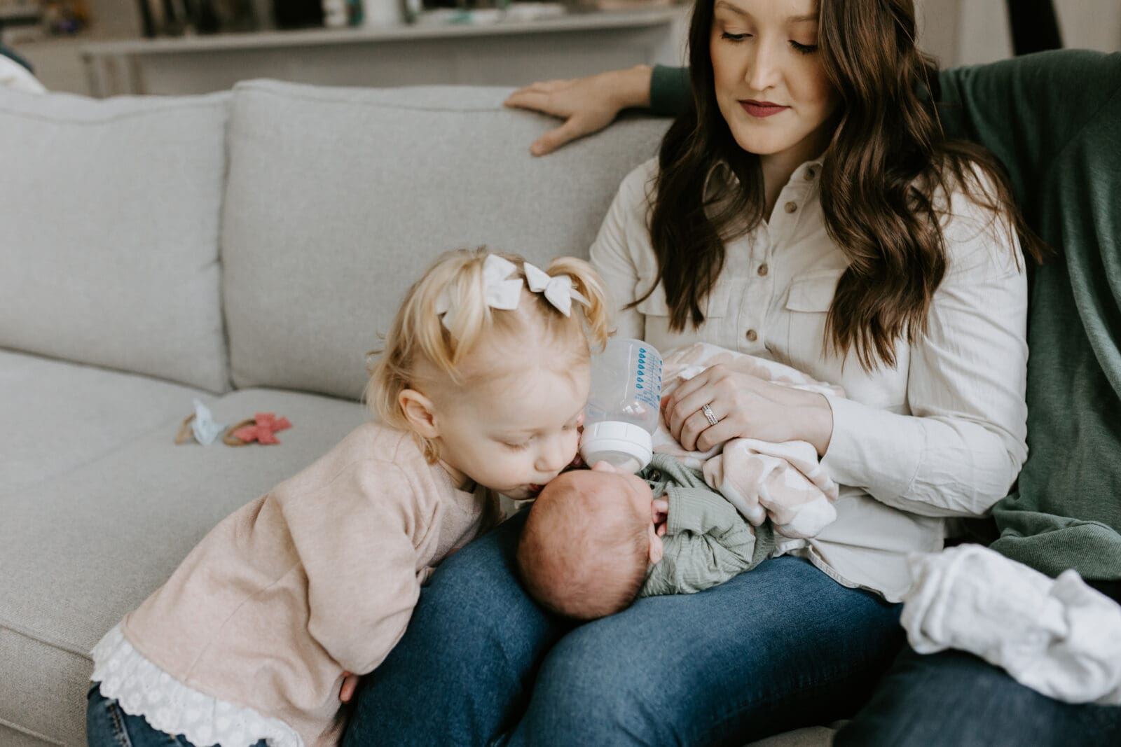 A mom feeds her newborn baby a bottle while an older sibling kisses the baby on the head.