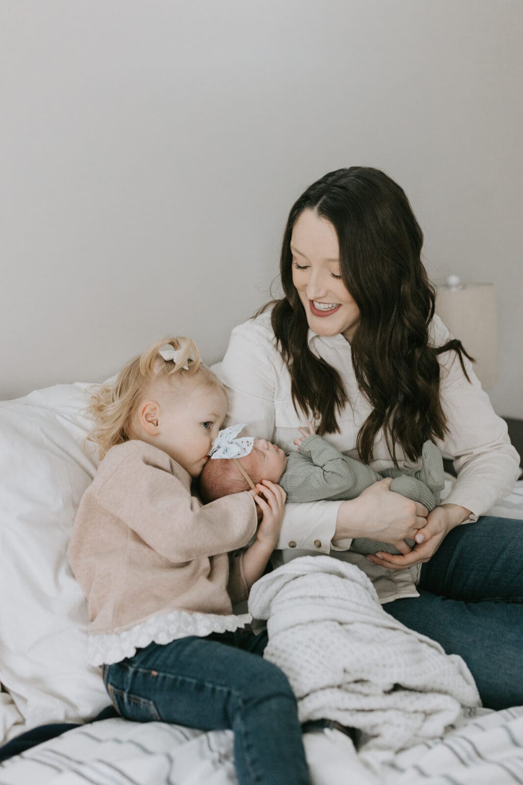 A mom holds a newborn baby on the bed while an older sibling kisses the baby on the head.