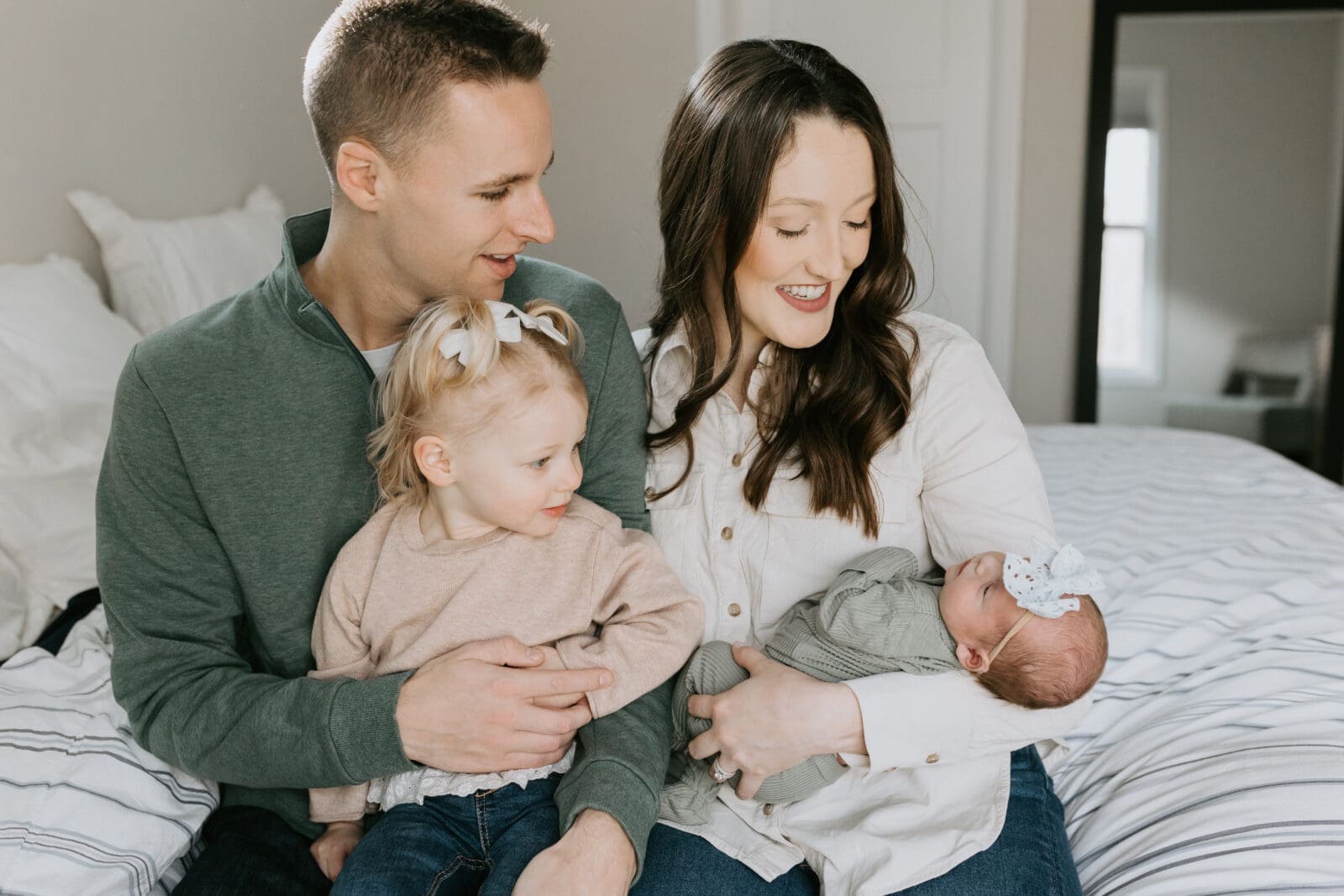 A mom, dad, and toddler smile at their newborn baby.