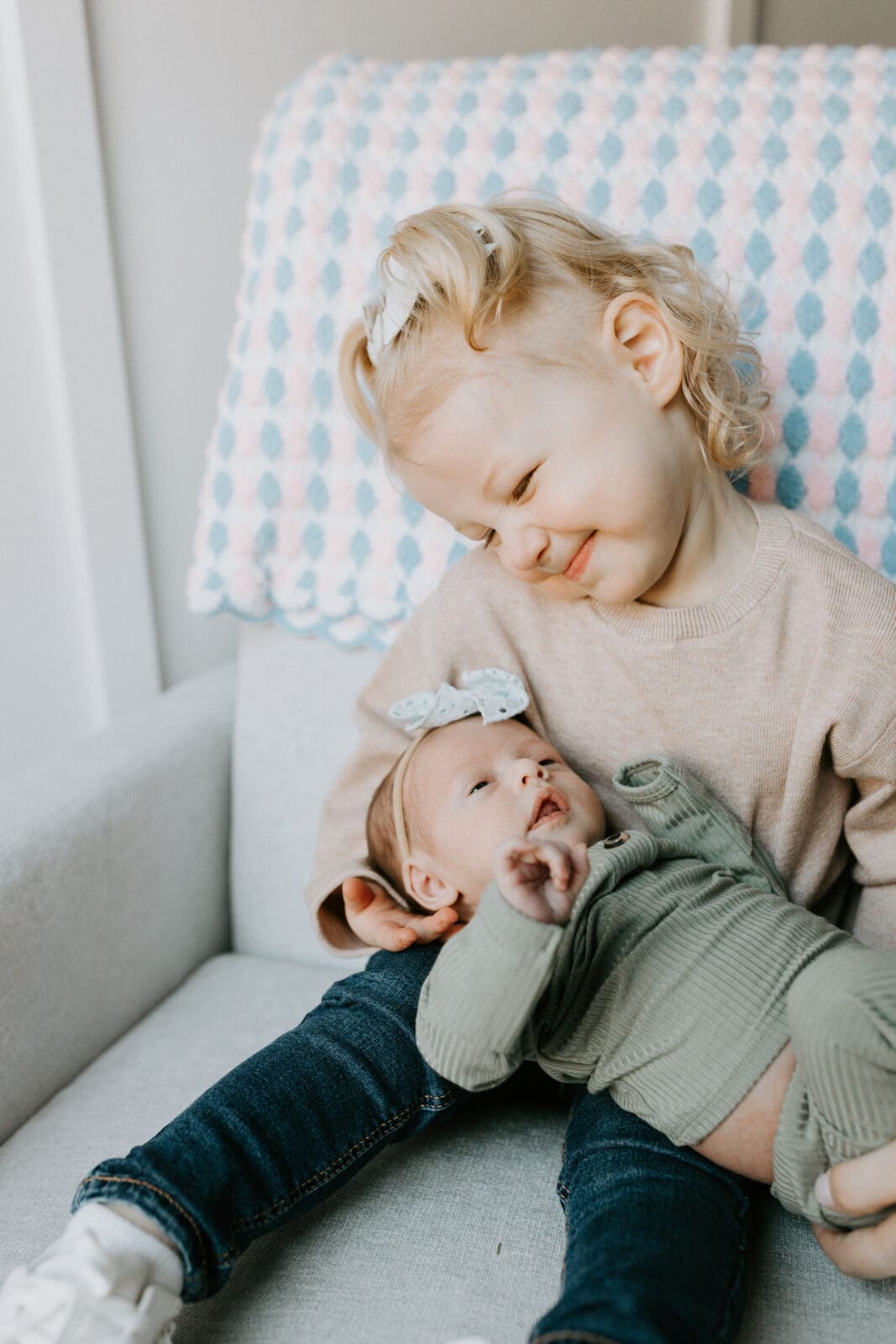 A toddler holds her baby sister on a rocking chair and smiles down at her.