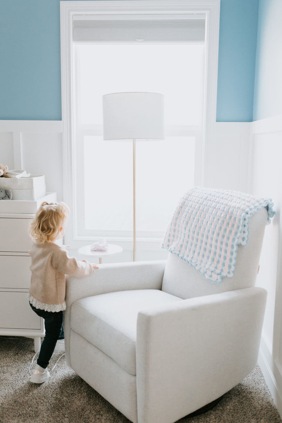 A toddler stands in a light blue and white nursery and looks out of the window.