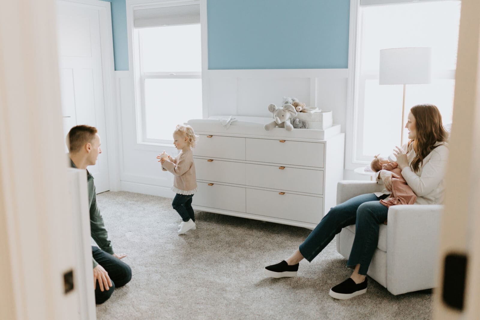 A mom sits on a recliner with her newborn baby in a nursery while her husband and toddler play together.
