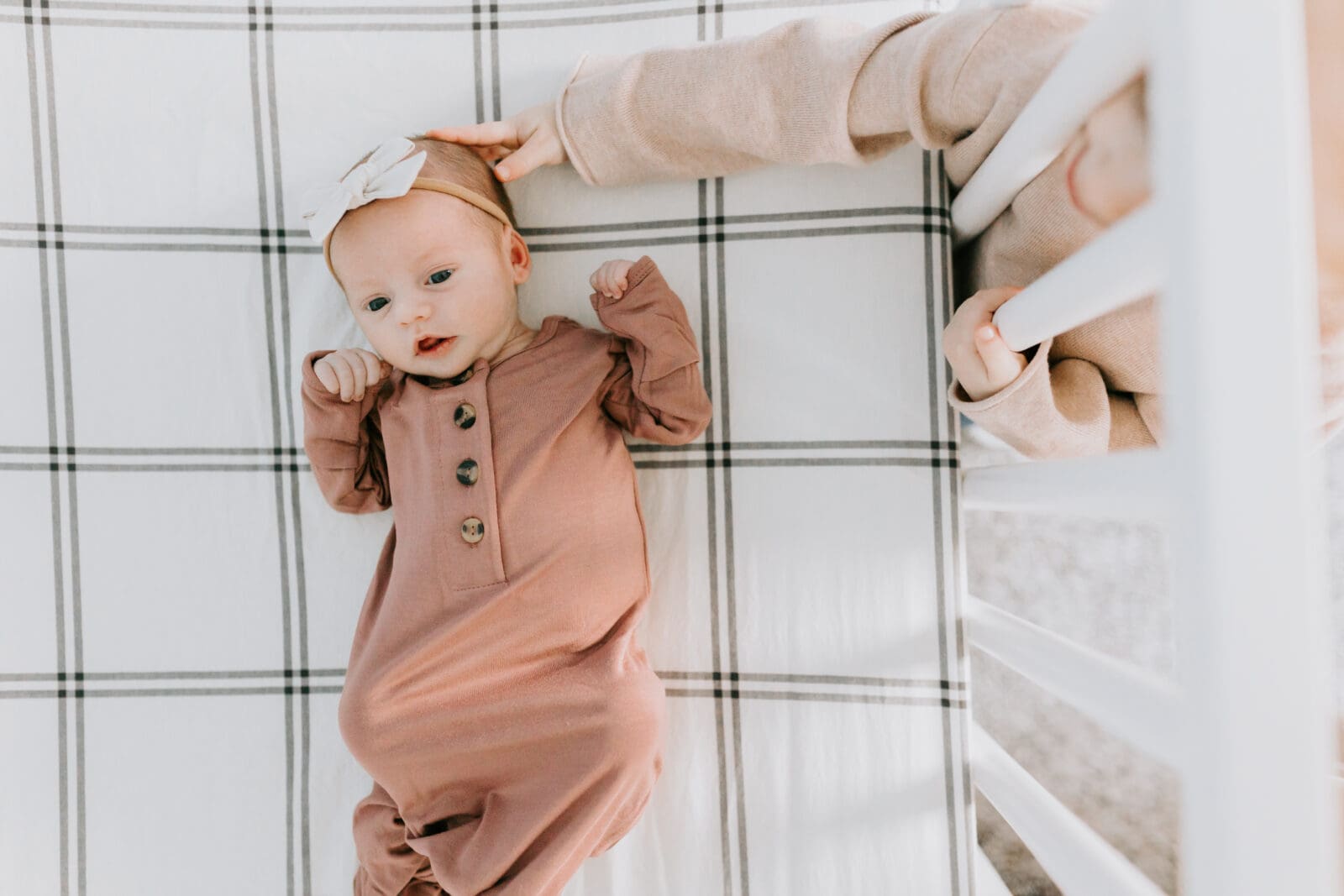 A toddler reaches her hand into a crib to gently touch the head of her newborn baby sister.